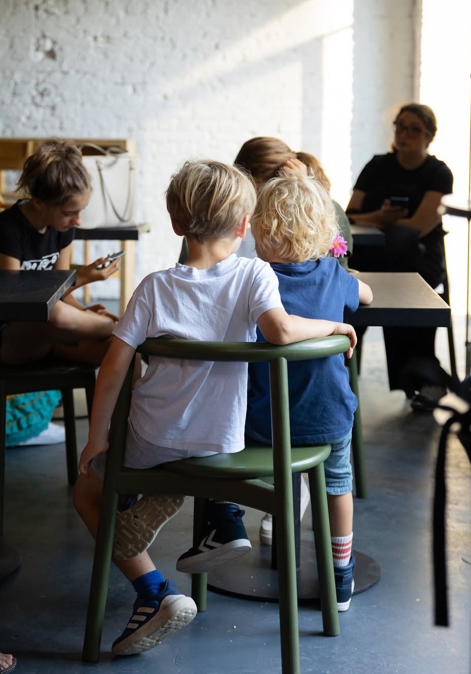 Zwei Kinder sitzen in einem Café auf Stühlen und blicken zur anderen Seite des Raums, umgeben von weiteren Gästen.
