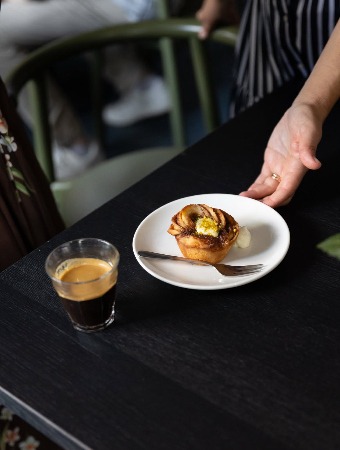 Eine Hand reicht einen Zimtschnecken-Muffin auf einem weißen Teller, daneben steht ein Glas Espresso auf einem schwarzen Tisch.