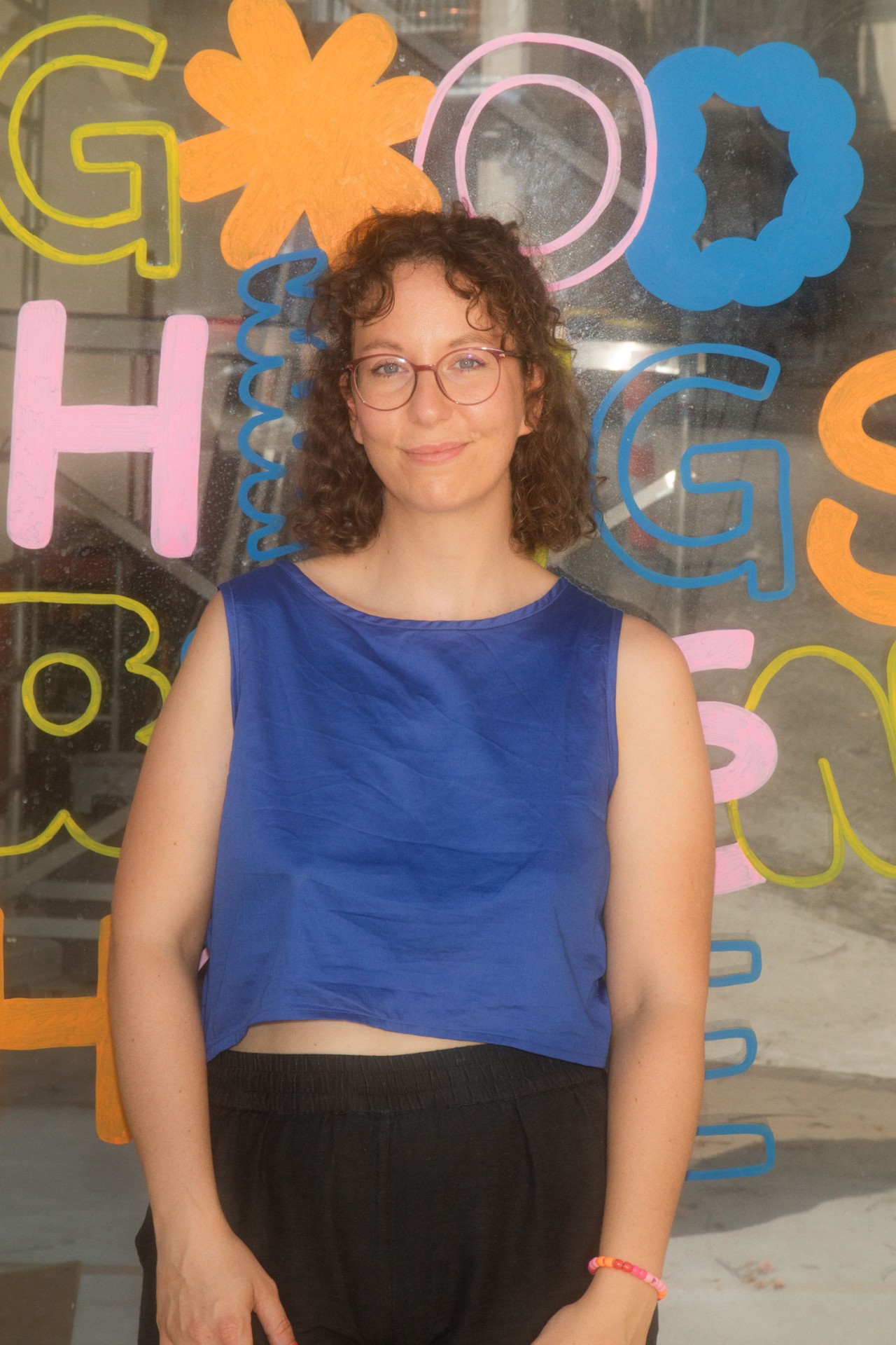 Frau mit lockigem Haar in blauer Bluse vor farbenfrohem Graffiti mit der Aufschrift „Good Things“.