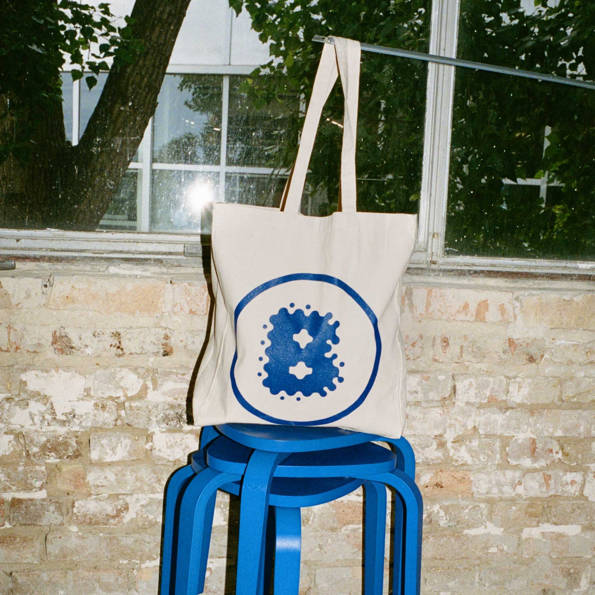Beige Baumwolltasche mit blauem Rundlogo auf einem blauen Hocker vor einem Fenster mit Pflanzen im Hintergrund.