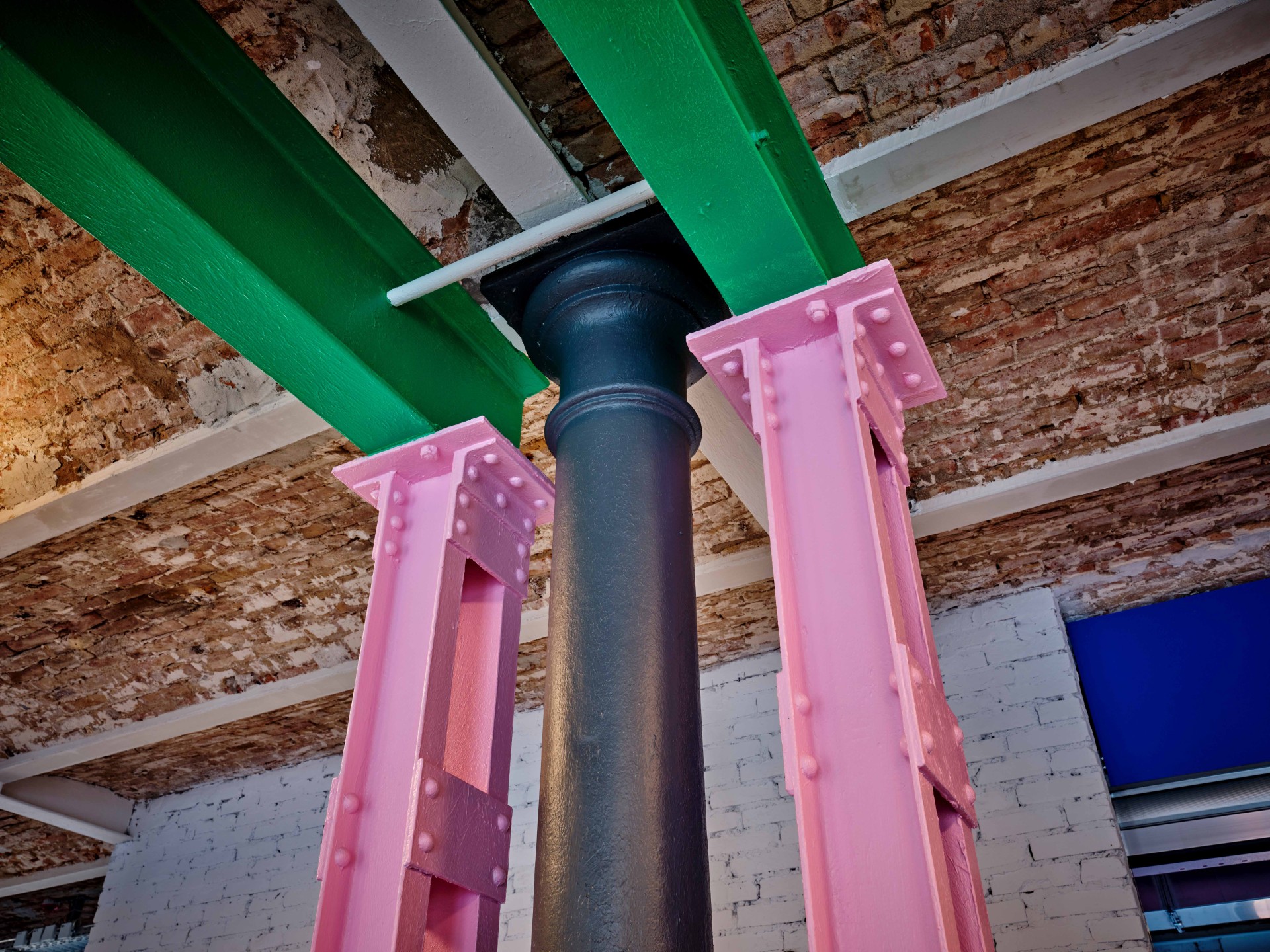 Bunt gestrichene Stahlträger in einem modernen Raum mit rohem Backstein und einem blauen Fenster im Hintergrund.