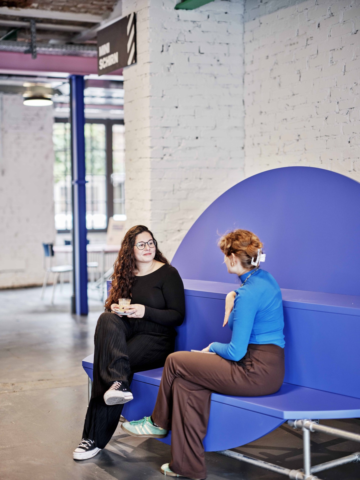 Zwei Frauen sitzen auf einer blauen Bank in einem modernen Raum und unterhalten sich, während eine von ihnen ein Getränk hält.