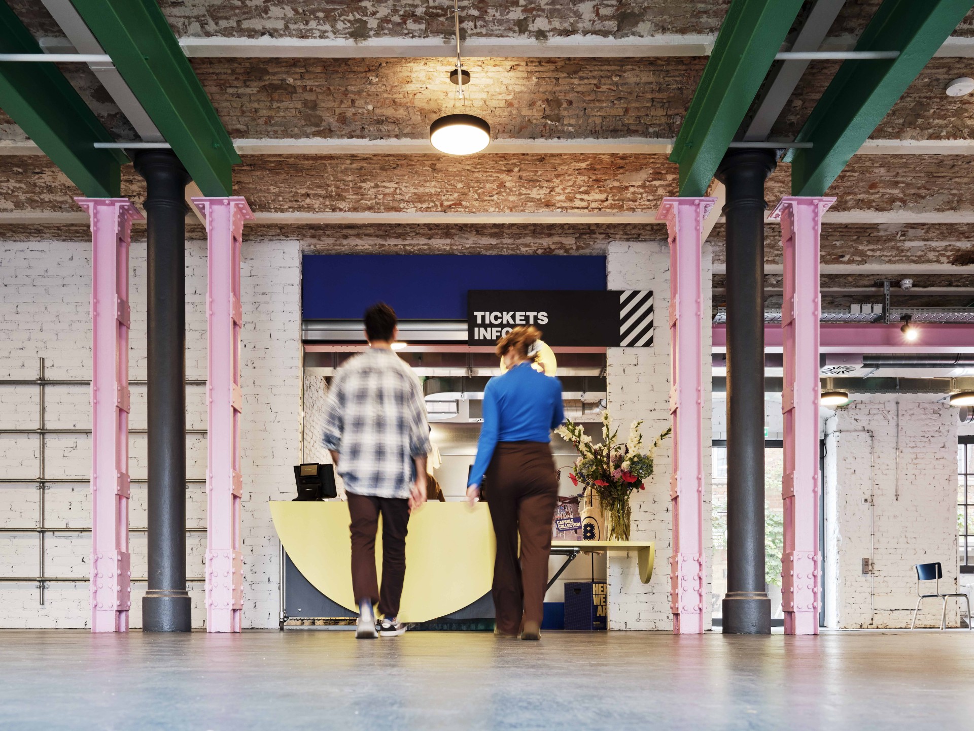Zwei Personen gehen in einem modernen Raum mit rosa und grünen Säulen zur Ticketinformation, im Hintergrund sind Blumen.
