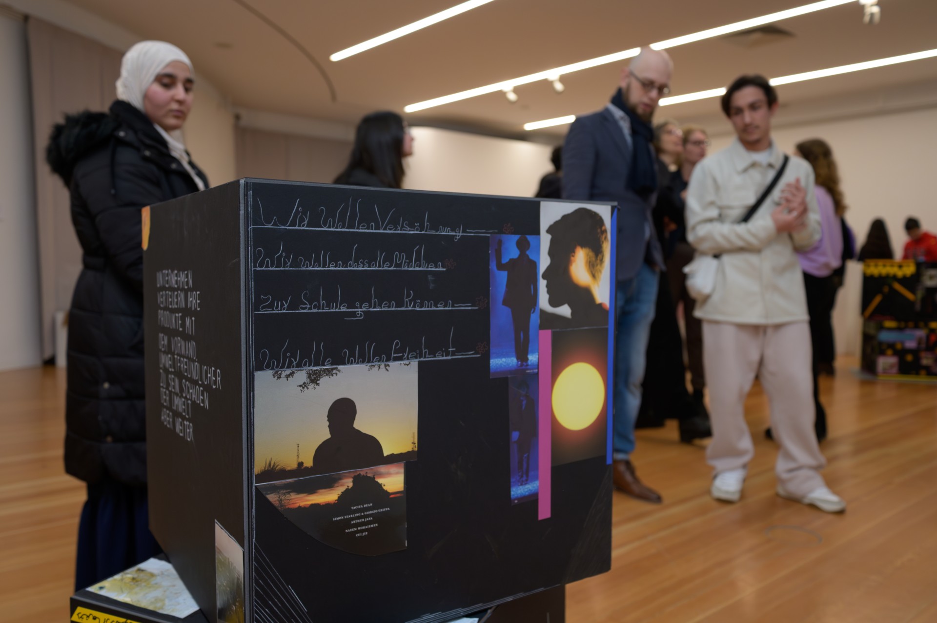 Ausstellungsszene mit mehreren Personen und einem künstlerisch gestalteten, informativen Objekt im Vordergrund.