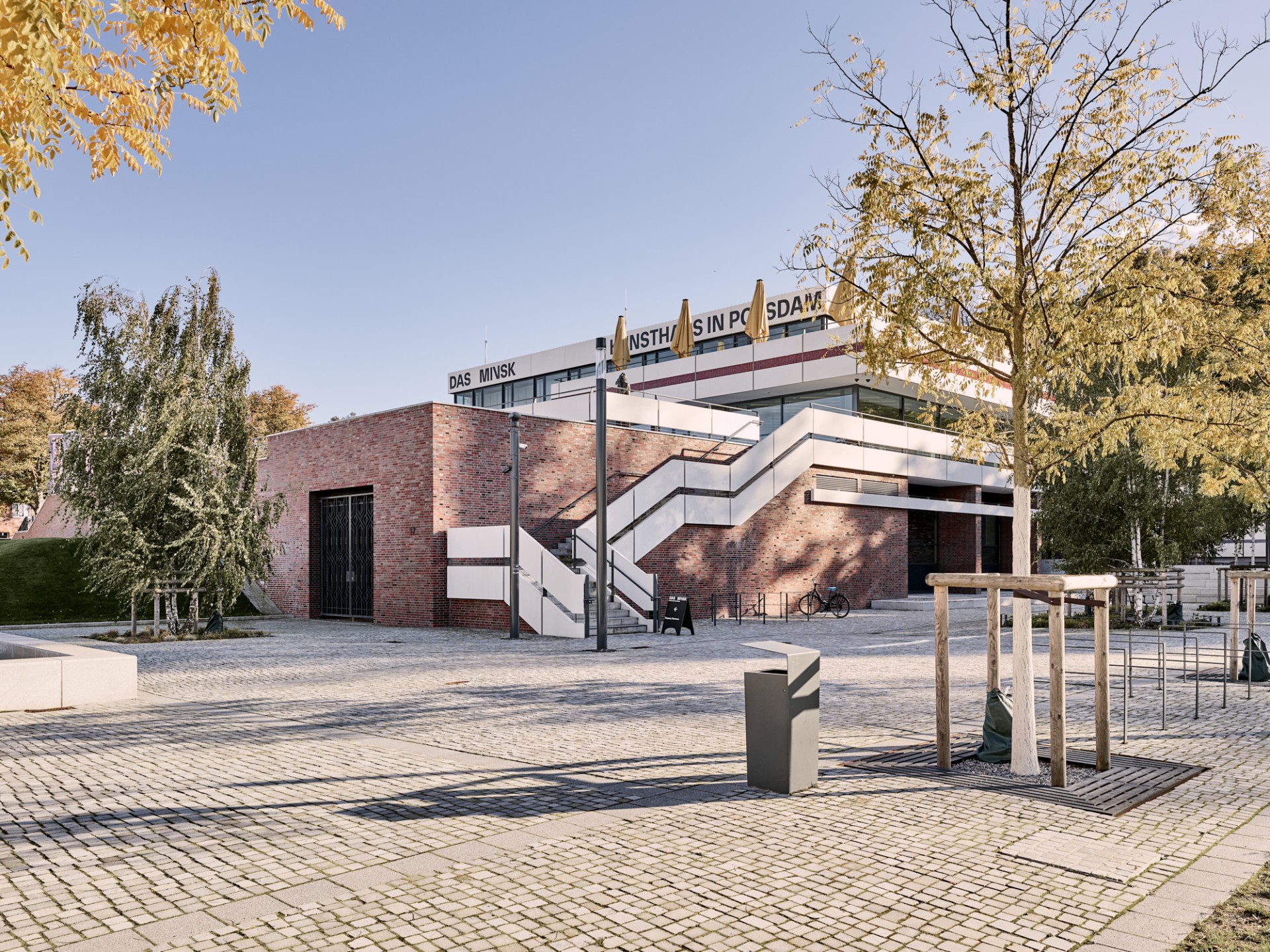 Modernes Gebäude in Potsdam mit Treppe, umgeben von Bäumen und Steinen, unter einem blauen Himmel.
