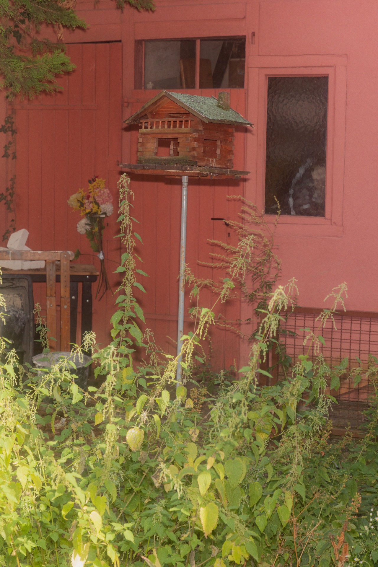 Ein nostalgisches Vogelhaus vor einer rosa Wand, umgeben von üppigen Pflanzen und einem bunten Blumenstrauß.