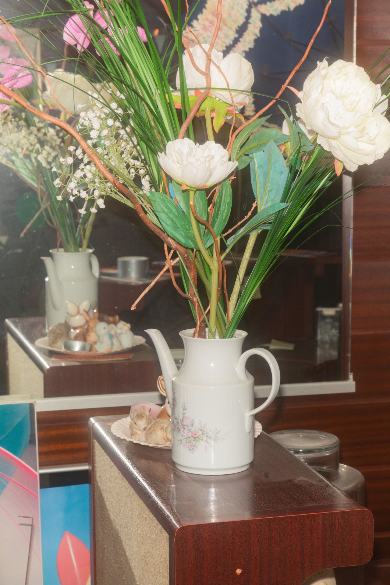 Kunstvolles Blumenarrangement in einer weißen Teekanne auf einem Holztisch, im Hintergrund ein Spiegel.