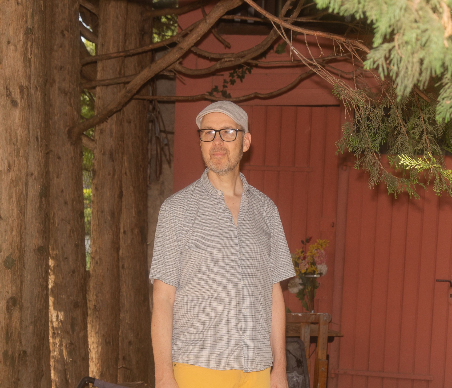 Mann im karierten Hemd und gelben Hosen steht im Schatten von Bäumen, umgeben von rustikaler Natur.