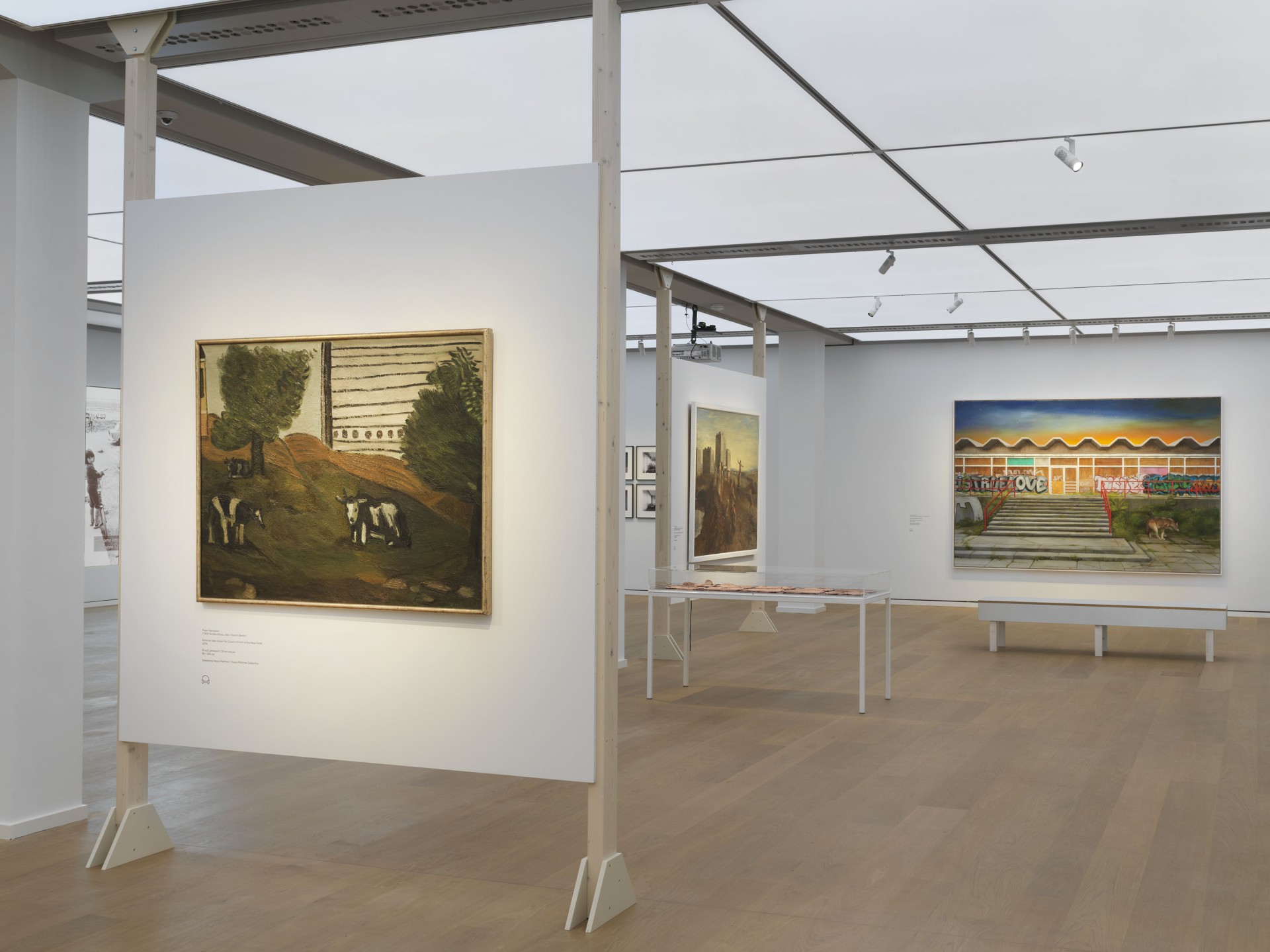 Moderne Galerie mit verschiedenen Gemälden, darunter Landschaften und urbane Kunst. Helle Wände und Holzfußboden.