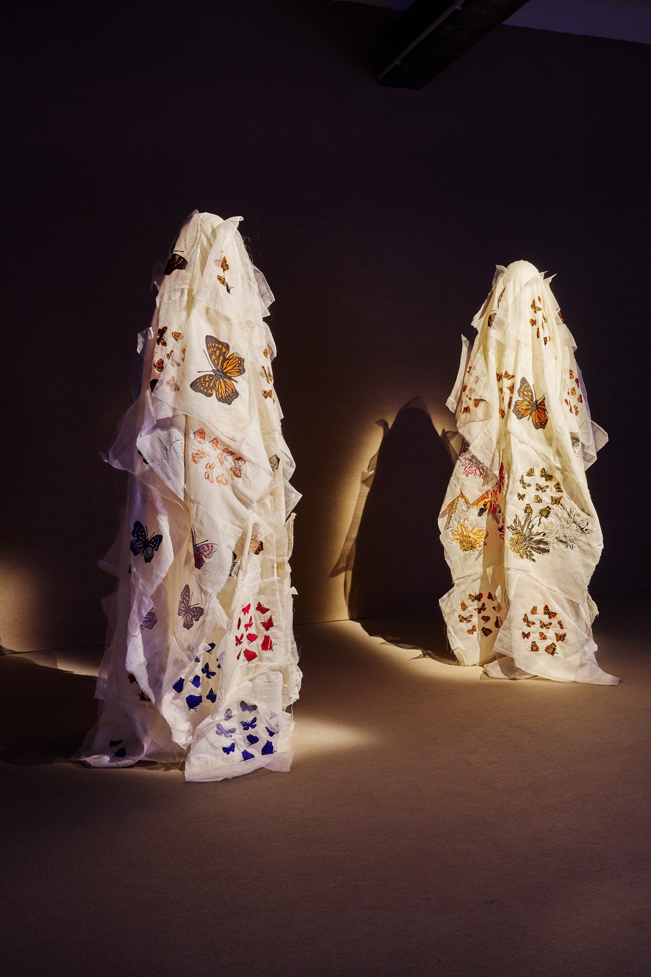 Two stylized fabric sculptures embroidered with butterflies stand in a lit room.