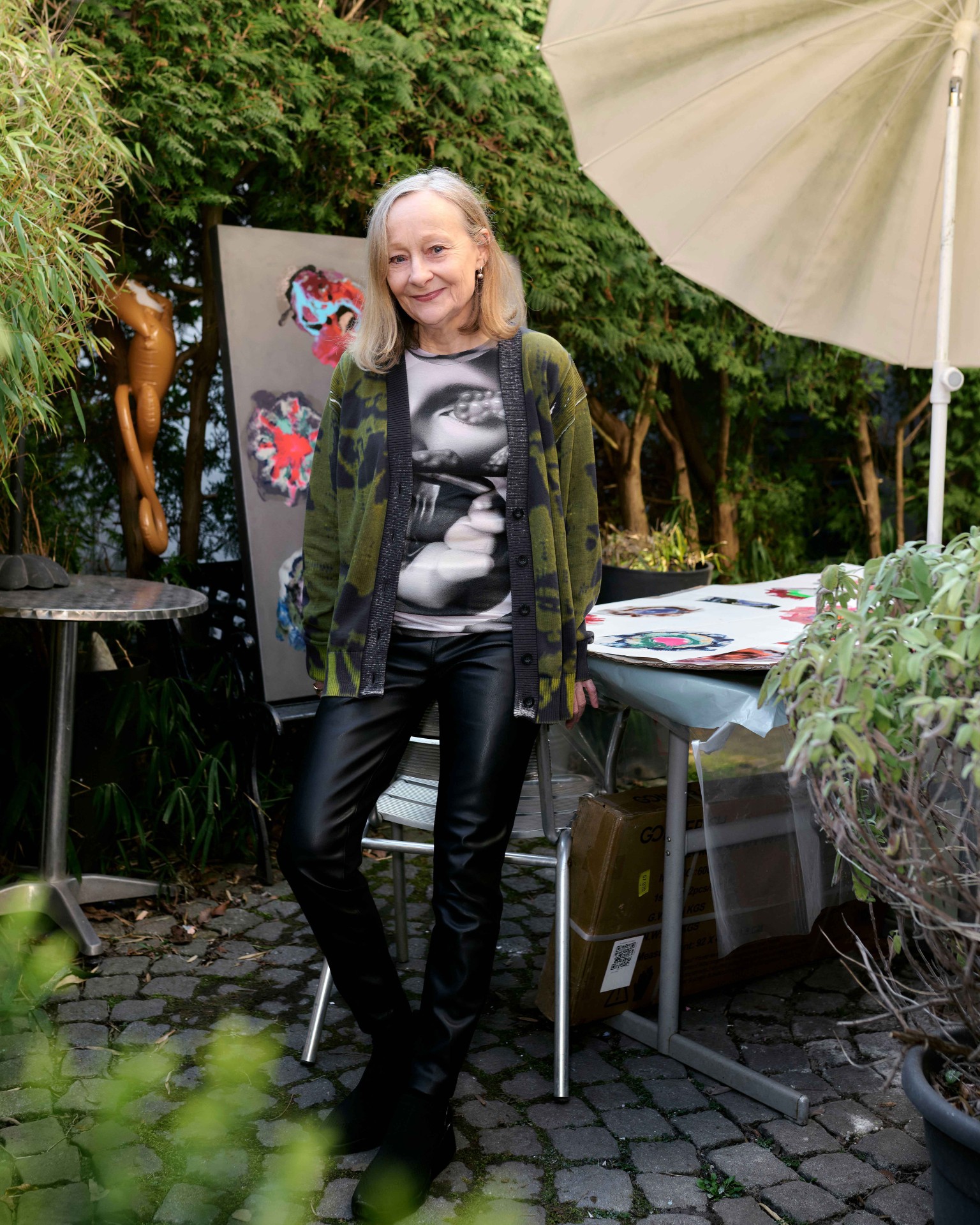 Frau mit langen, grauen Haaren im gestreiften Cardigan steht im Garten vor einem Tisch mit Kunstwerken.
