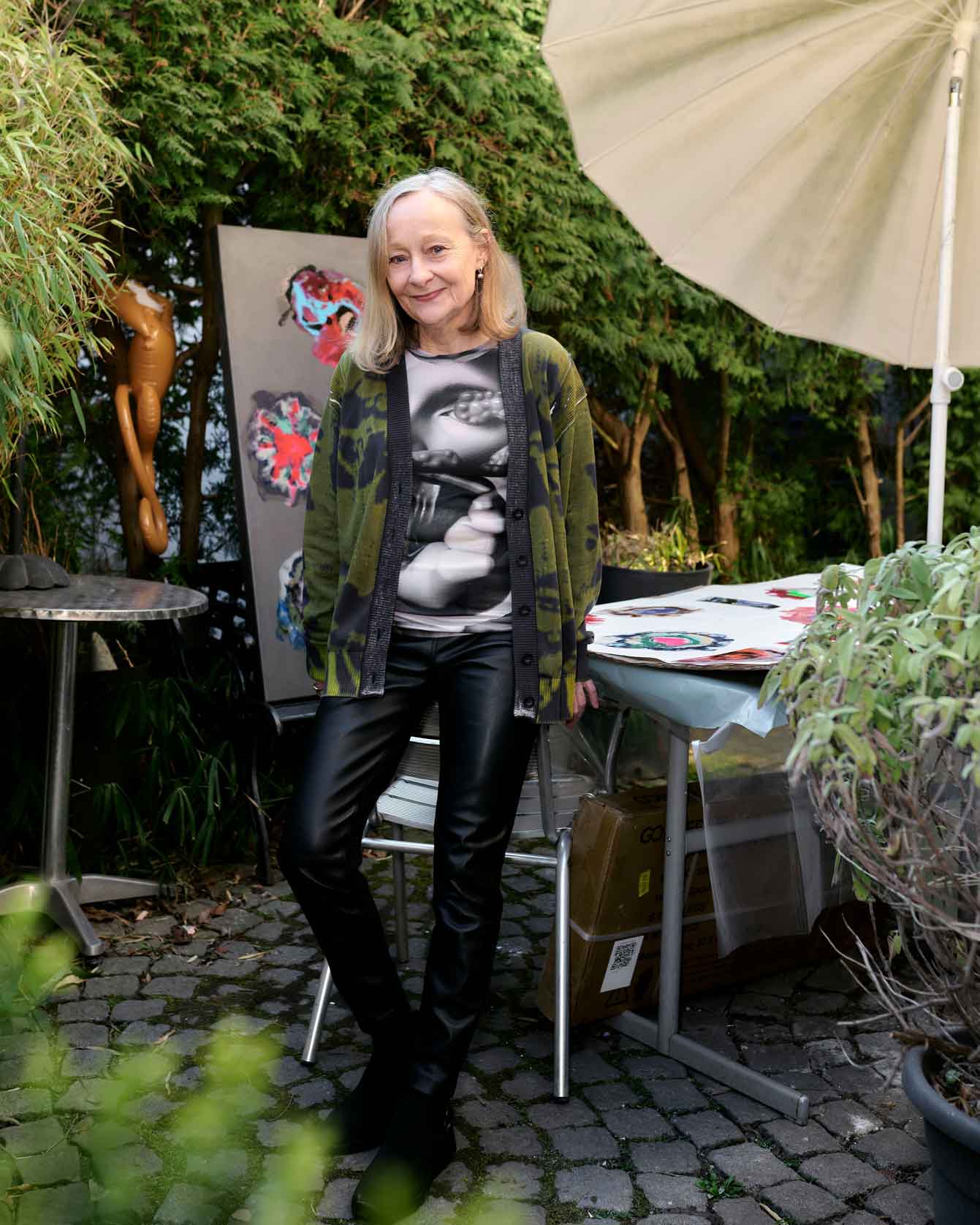 Eine lächelnde Frau in einem natürlichen Garten trägt eine bunte Jacke und schwarze Hosen, umgeben von Kunstwerken.