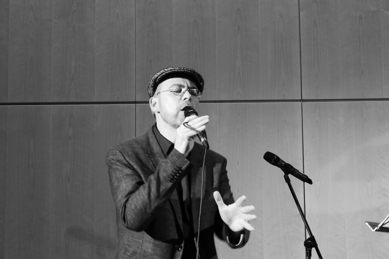 Ein Mann singt leidenschaftlich ins Mikrofon, gekleidet in Anzug und Mütze, vor einer Wand aus Holzpaneelen.