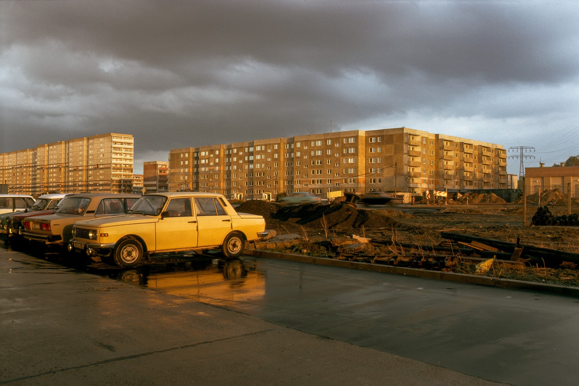 Gelbe Oldtimer stehen vor grauen Plattenbauten, der Himmel ist bewölkt, und das Licht reflektiert auf dem Asphalt.