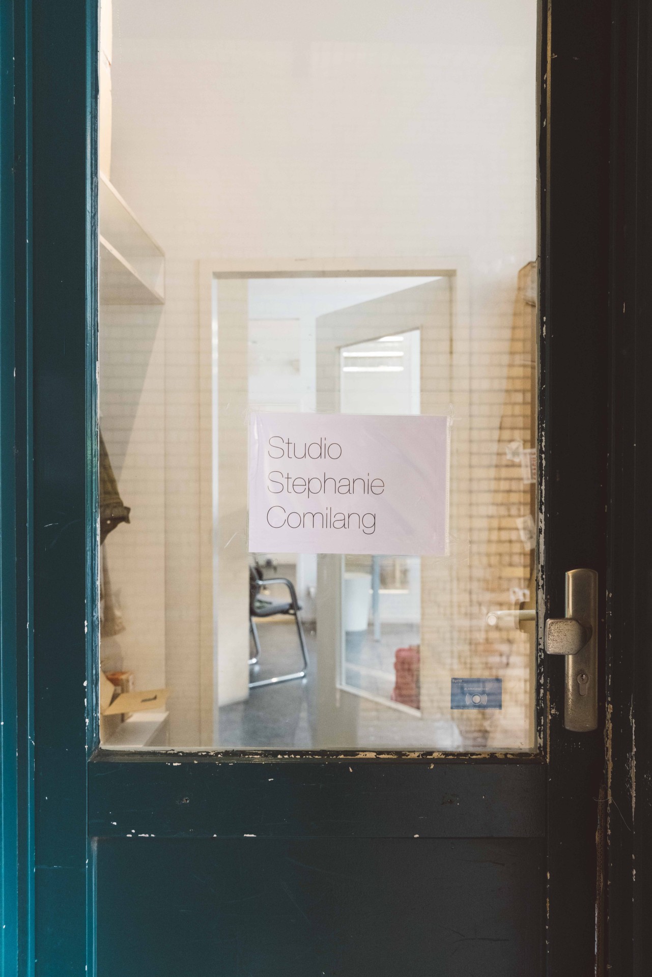 Door with a glass window and a sign saying "Studio Stephanie Comilang," with a bright room in the background.