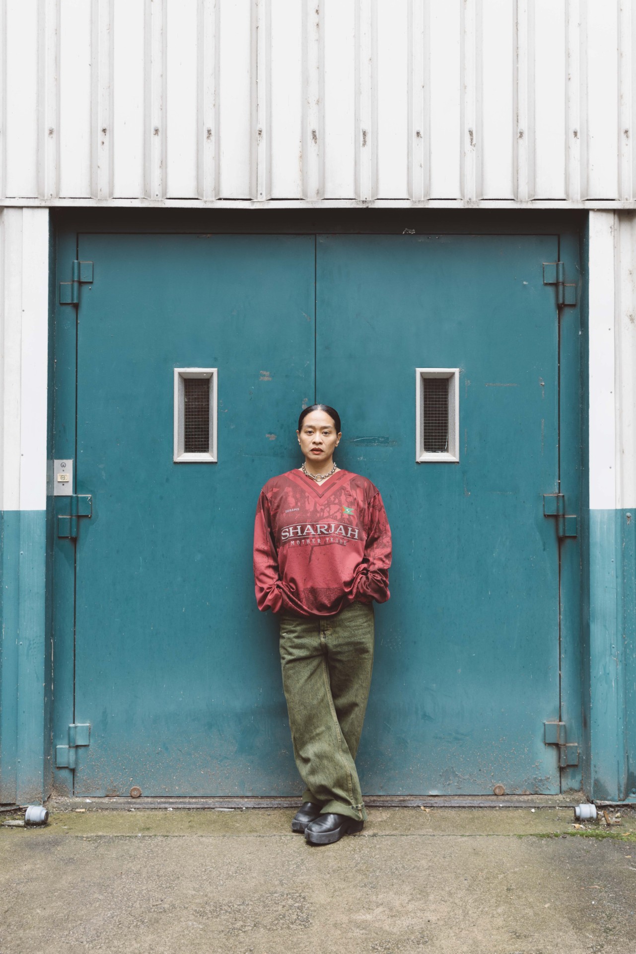 A young person in a red sweatshirt and green pants leans dreamily against a blue door.