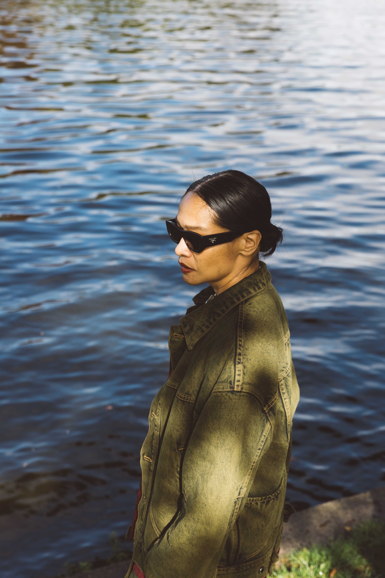 A woman in sunglasses and a green jacket stands by the water, thoughtfully gazing into the distance.