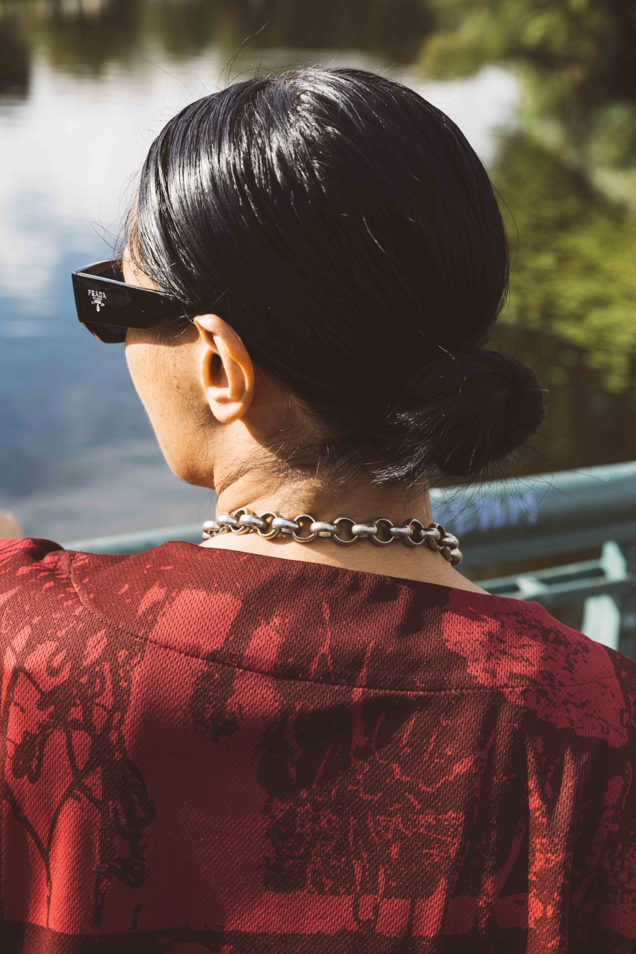 Woman in sunglasses with a chain, photographed from behind, by the shore of a body of water with lush vegetation.