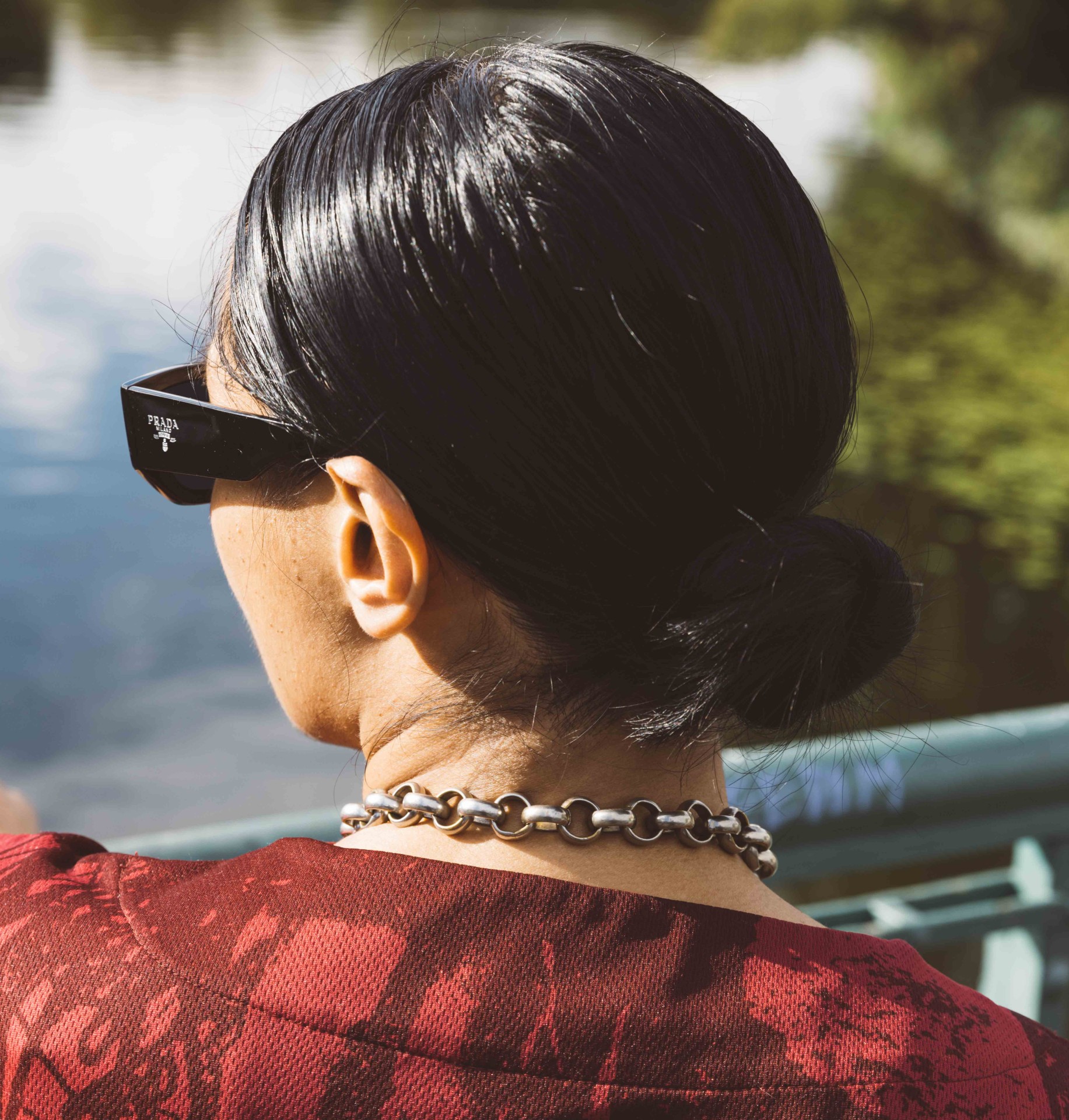 Frau mit Sonnenbrille und Kette, von hinten am Ufer eines Gewässers mit üppiger Vegetation fotografiert.