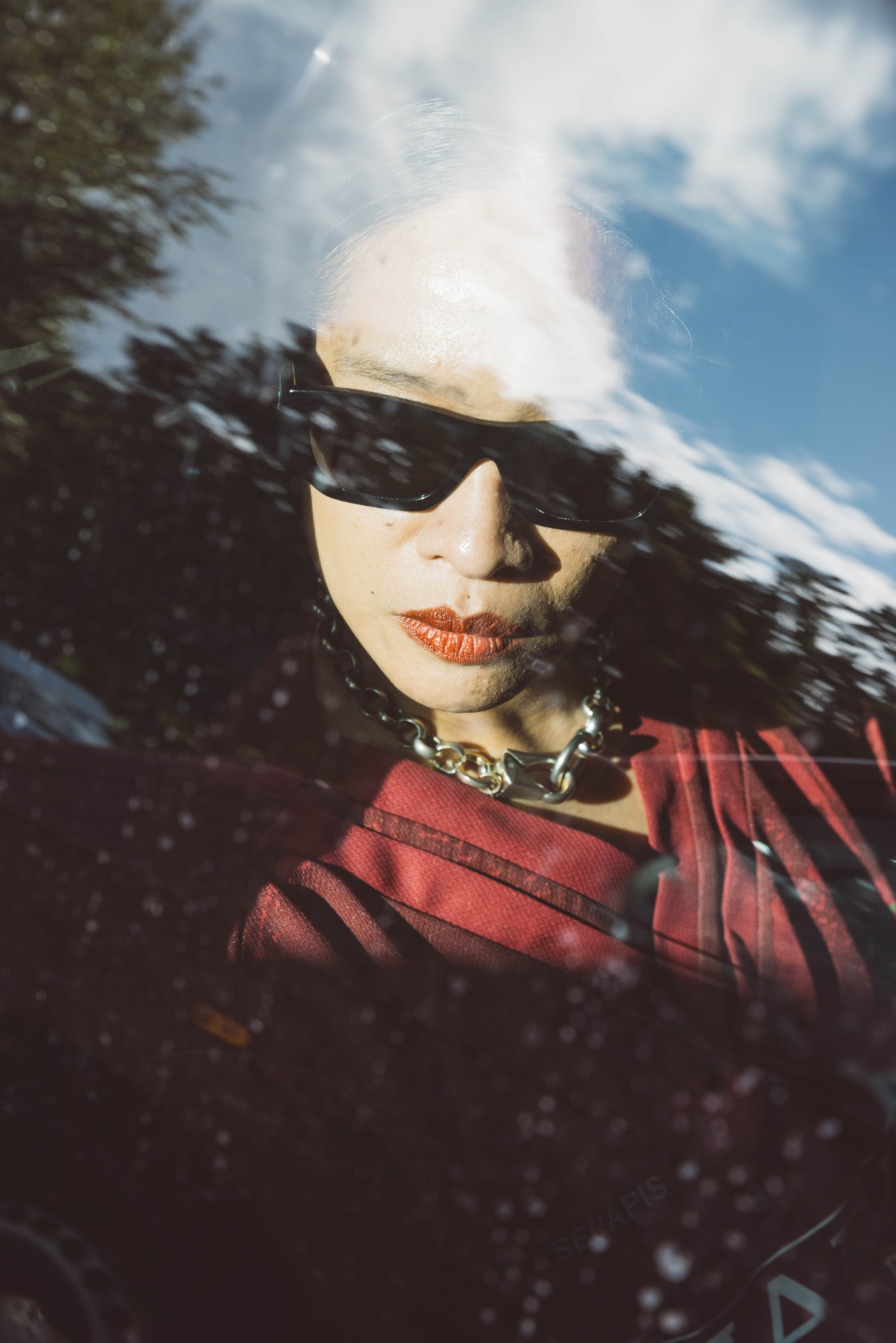 Reflection of a person wearing sunglasses and a red top, surrounded by nature and clouds.