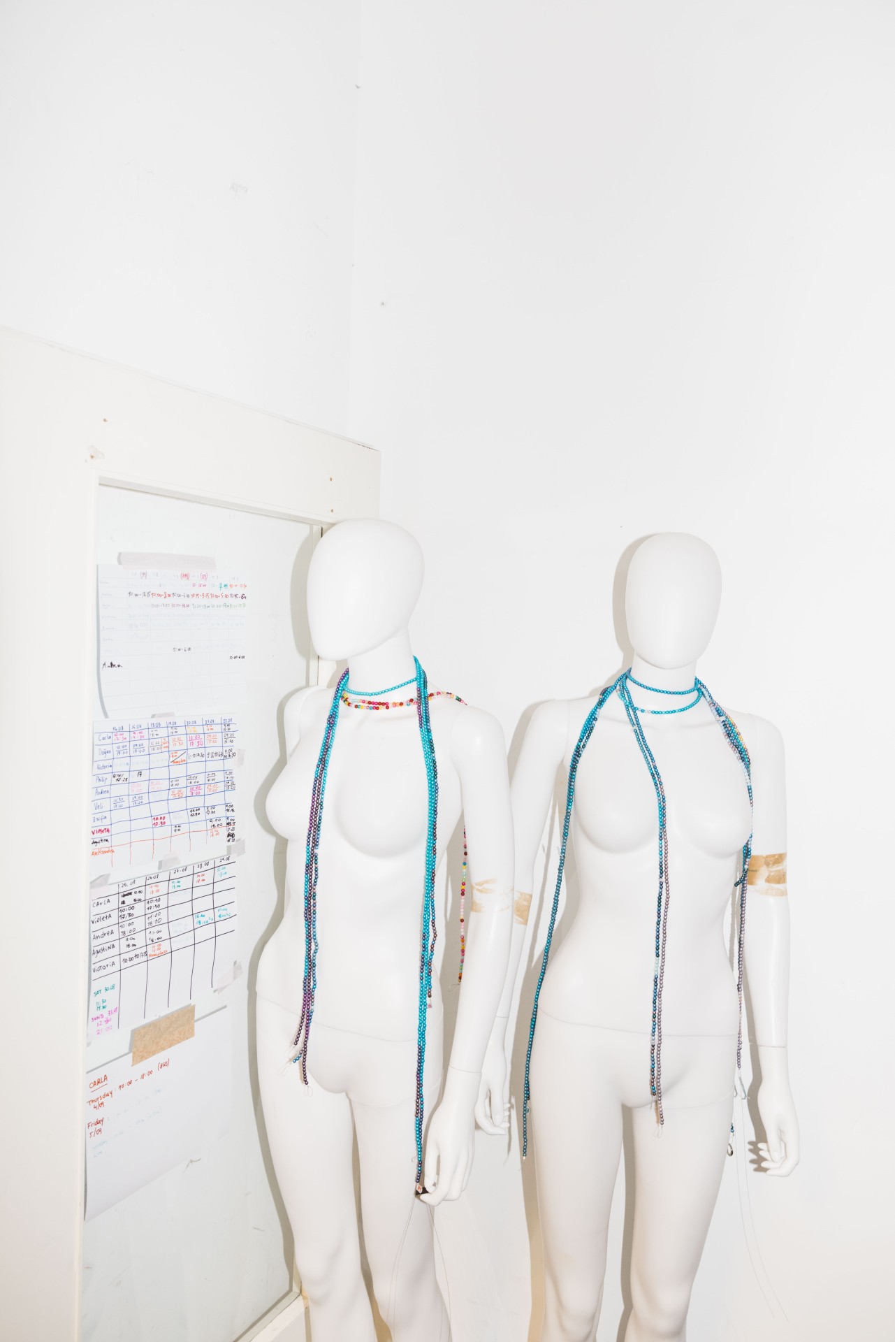Two white mannequins with colorful necklaces in front of a white wall and an organizational board in the background.