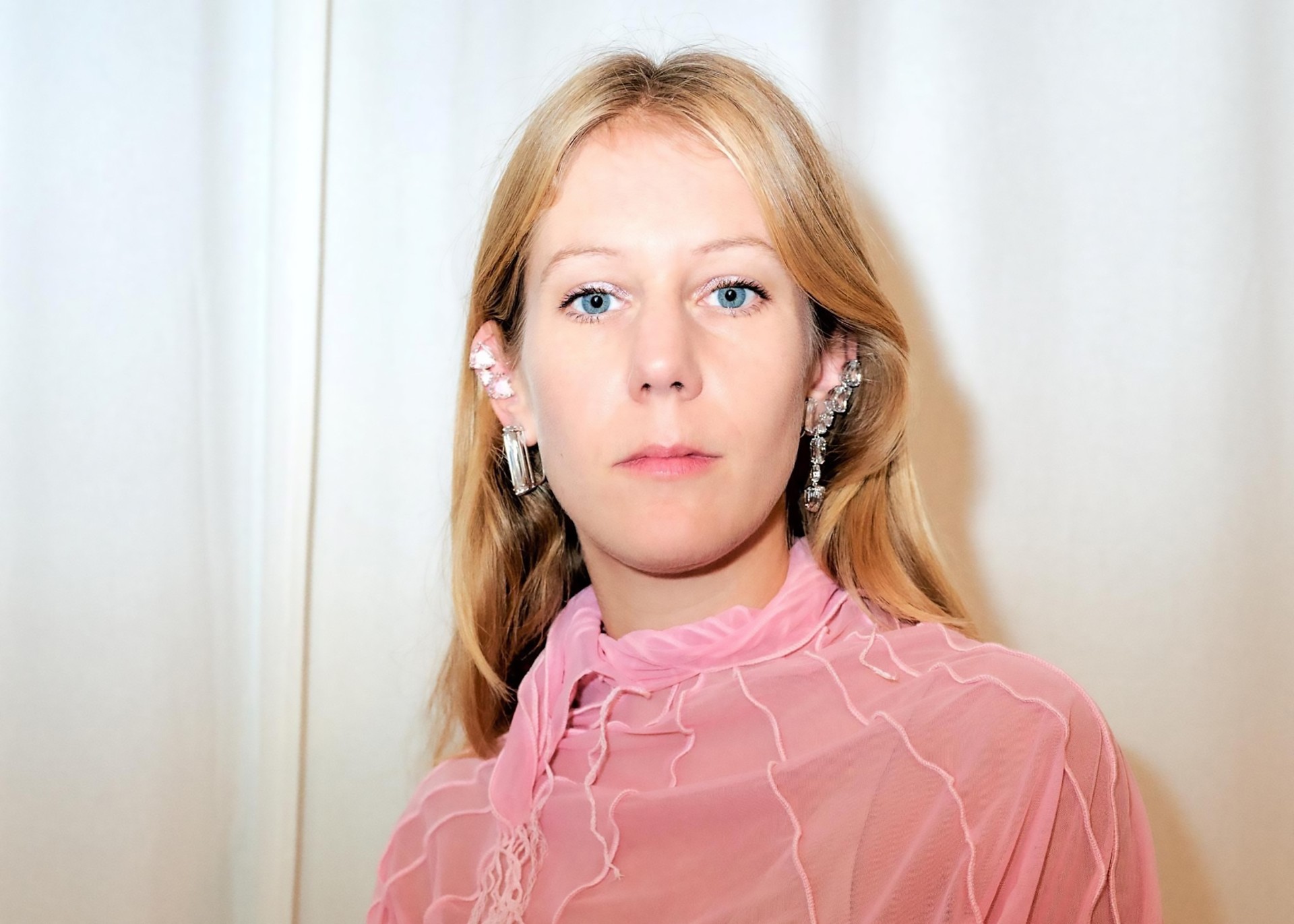 Woman with long blonde hair, blue eyes, in a pink top and striking earrings against a light background.