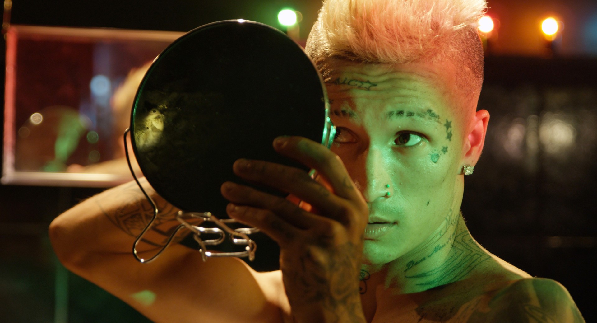 Eine Person mit Tattoos und blonden Haaren blickt in einen Spiegel, umgeben von bunten Lichtern.