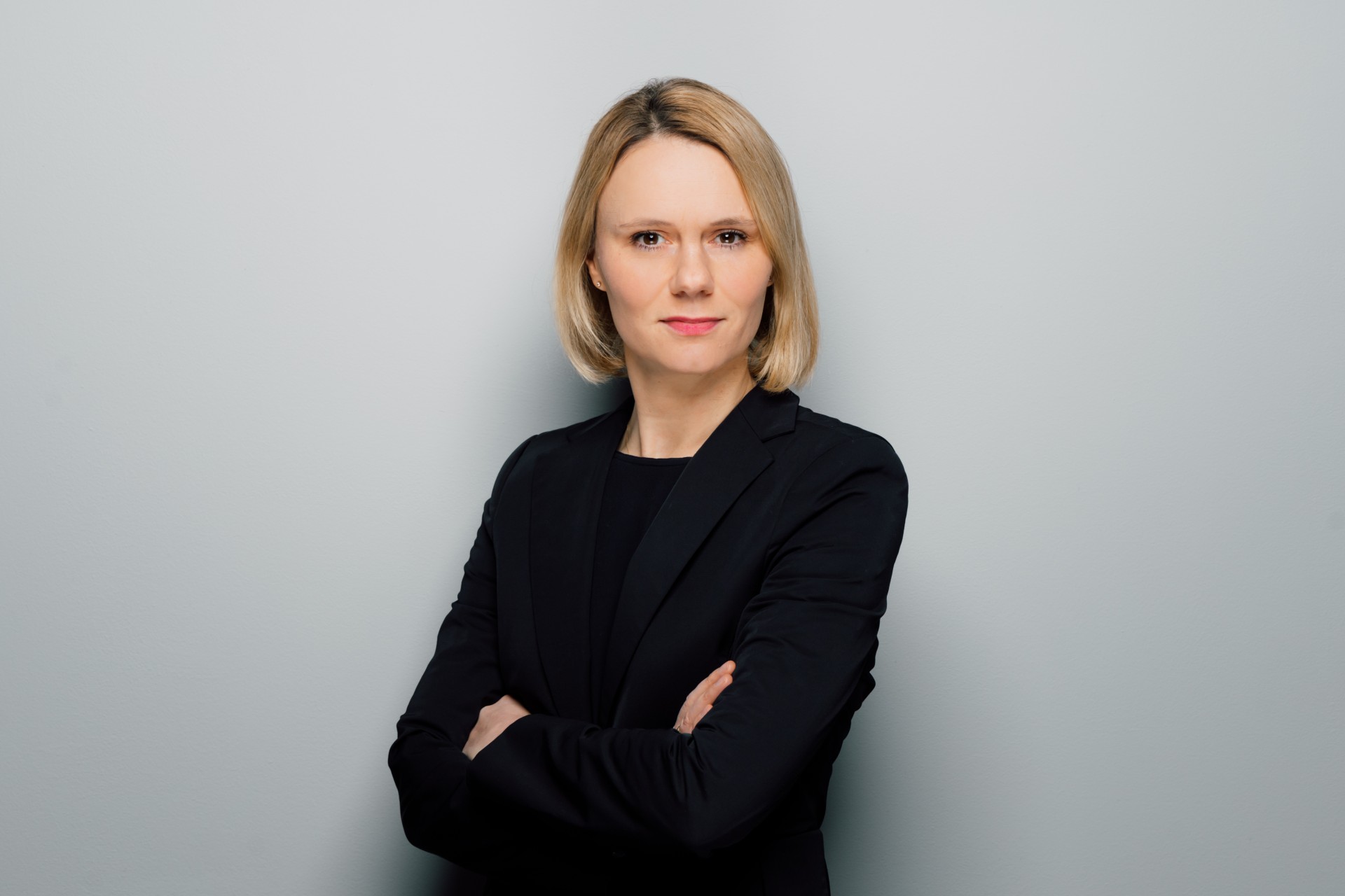 Portrait of a confident woman in a black blazer with crossed arms against a gray background.