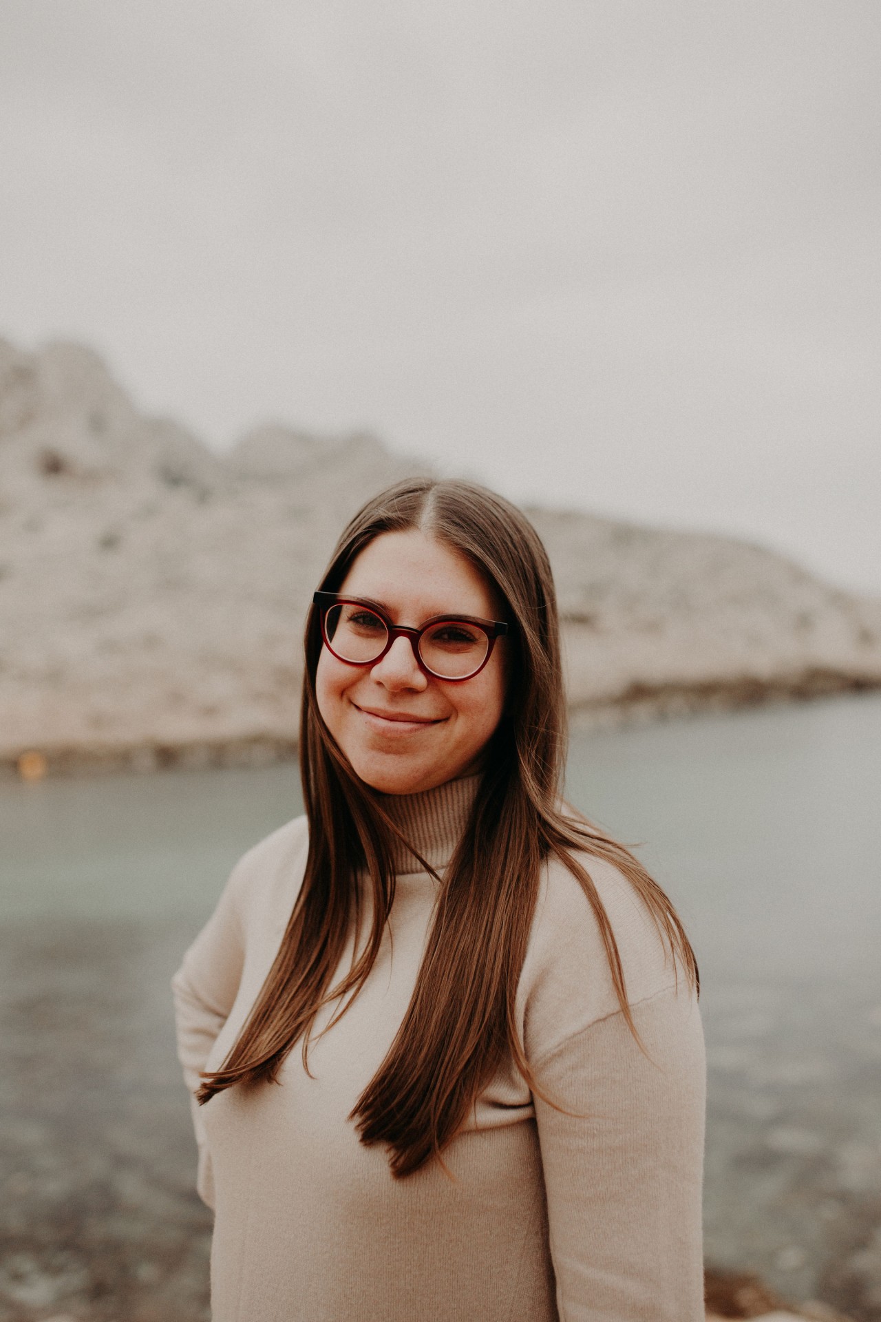 Porträt einer lächelnden Frau mit langen Haaren und Brille, stehend am Wasser mit Felsen im Hintergrund.