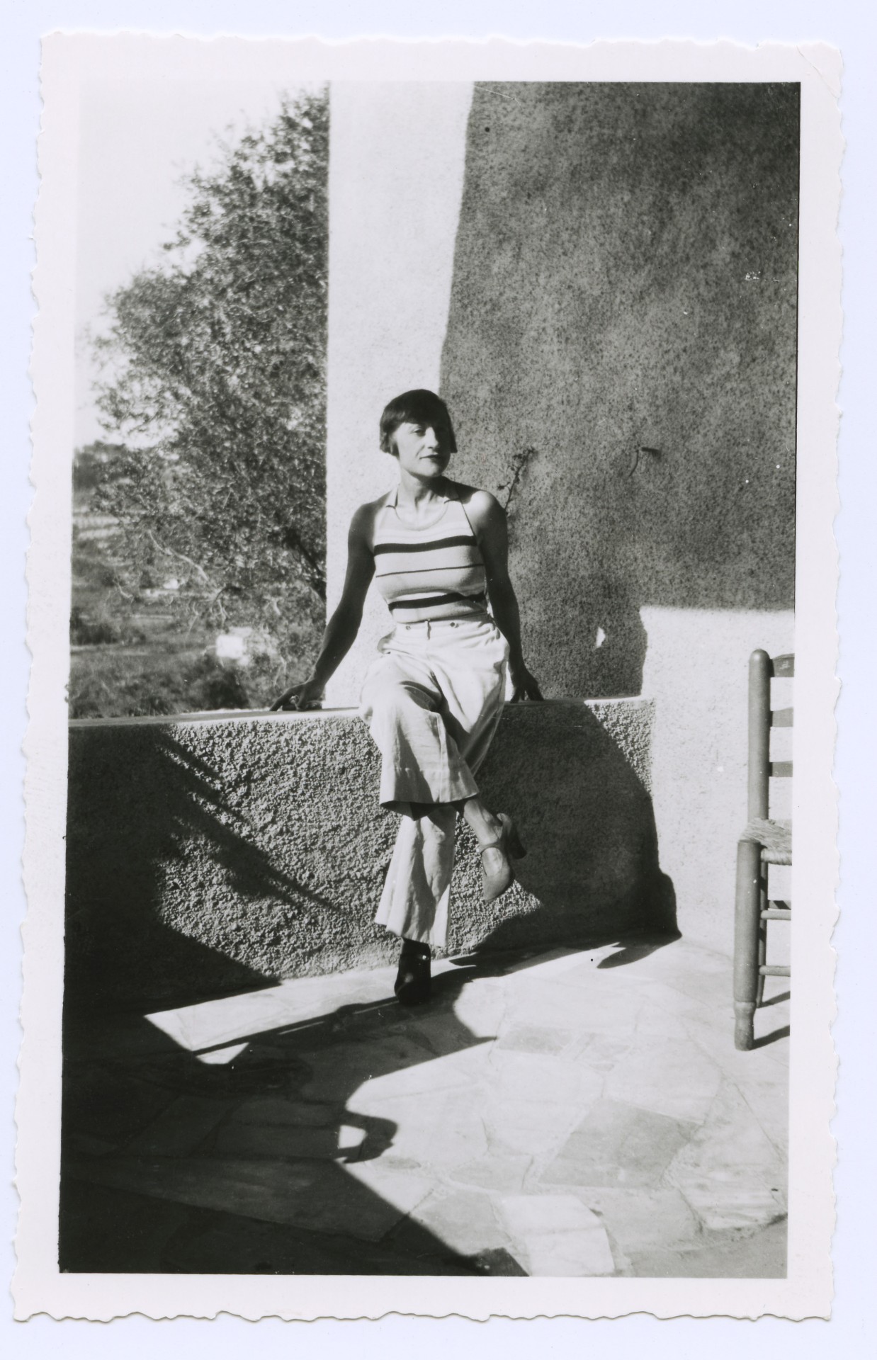 Eine Frau sitzt entspannt auf einer Mauer, trägt ein gestreiftes Top und weite Hosen, umgeben von Natur.