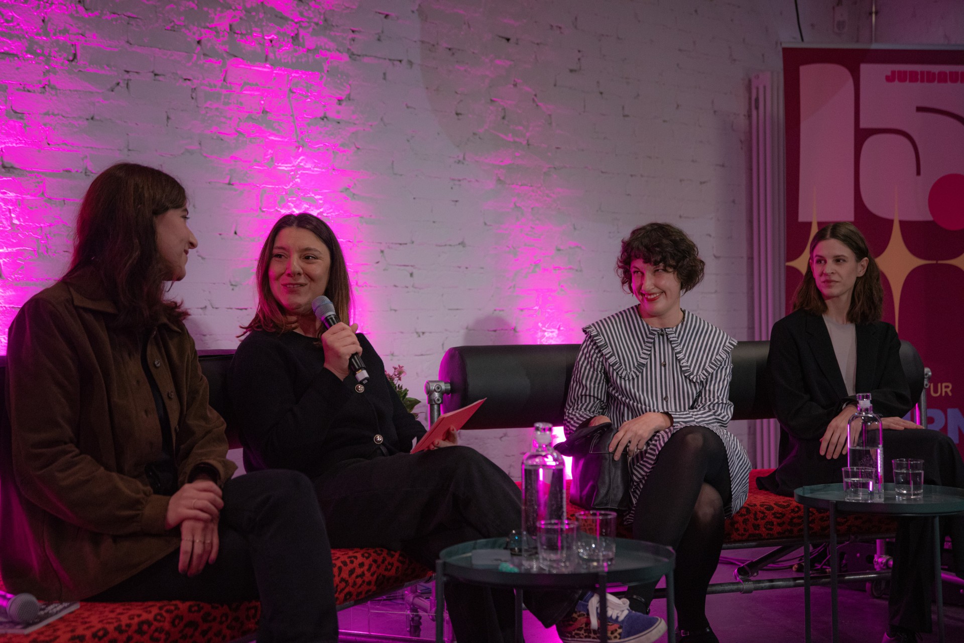Podiumsdiskussion mit vier Frauen auf Sofas, vor einer pinken Lichterwand und mit Mikrofonen in der Hand.