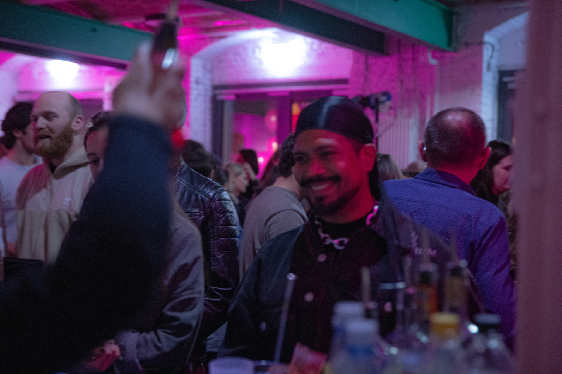 Menschenmenge bei einem Event, mit einem lächelnden Mann in dunkler Kleidung, umgeben von bunten Lichtern im Fokus.