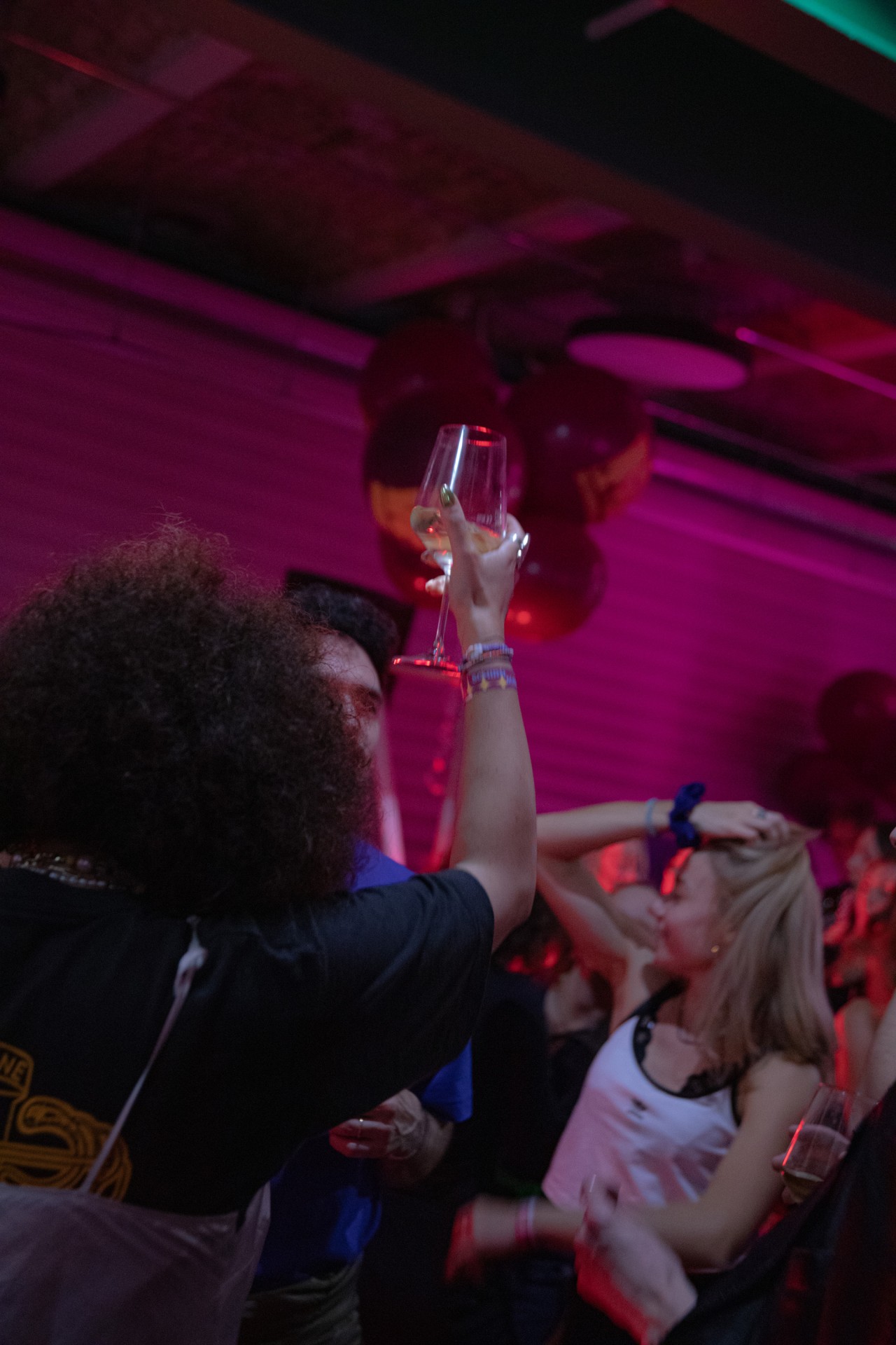 Menschen feiern in einer lebhaften Atmosphäre mit Getränken, pinkem Licht und Luftballons.