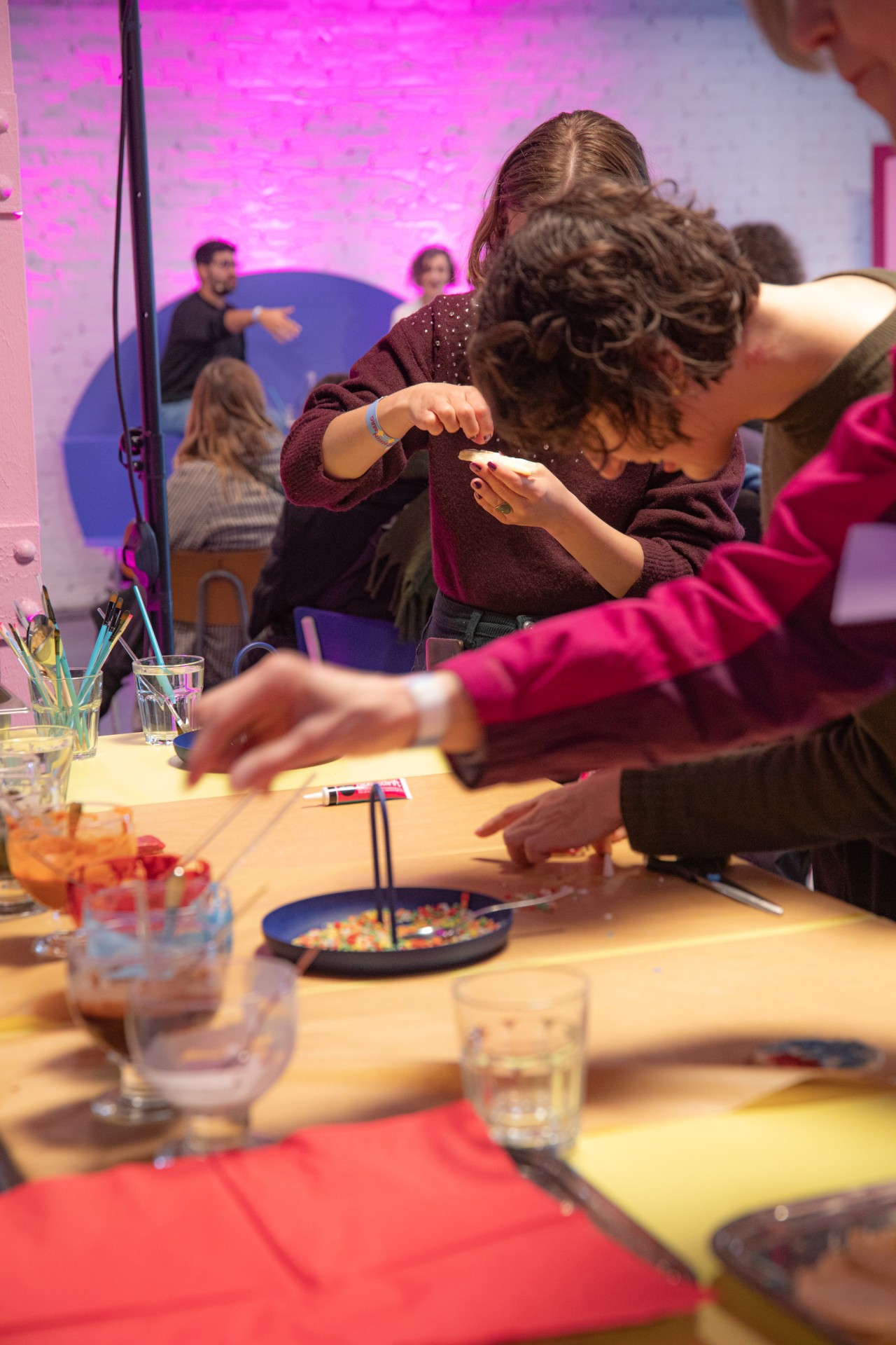 Tisch mit Getränken, Materialien und Menschen, die kreativ arbeiten und dekorieren, vor einem bunten Hintergrund.