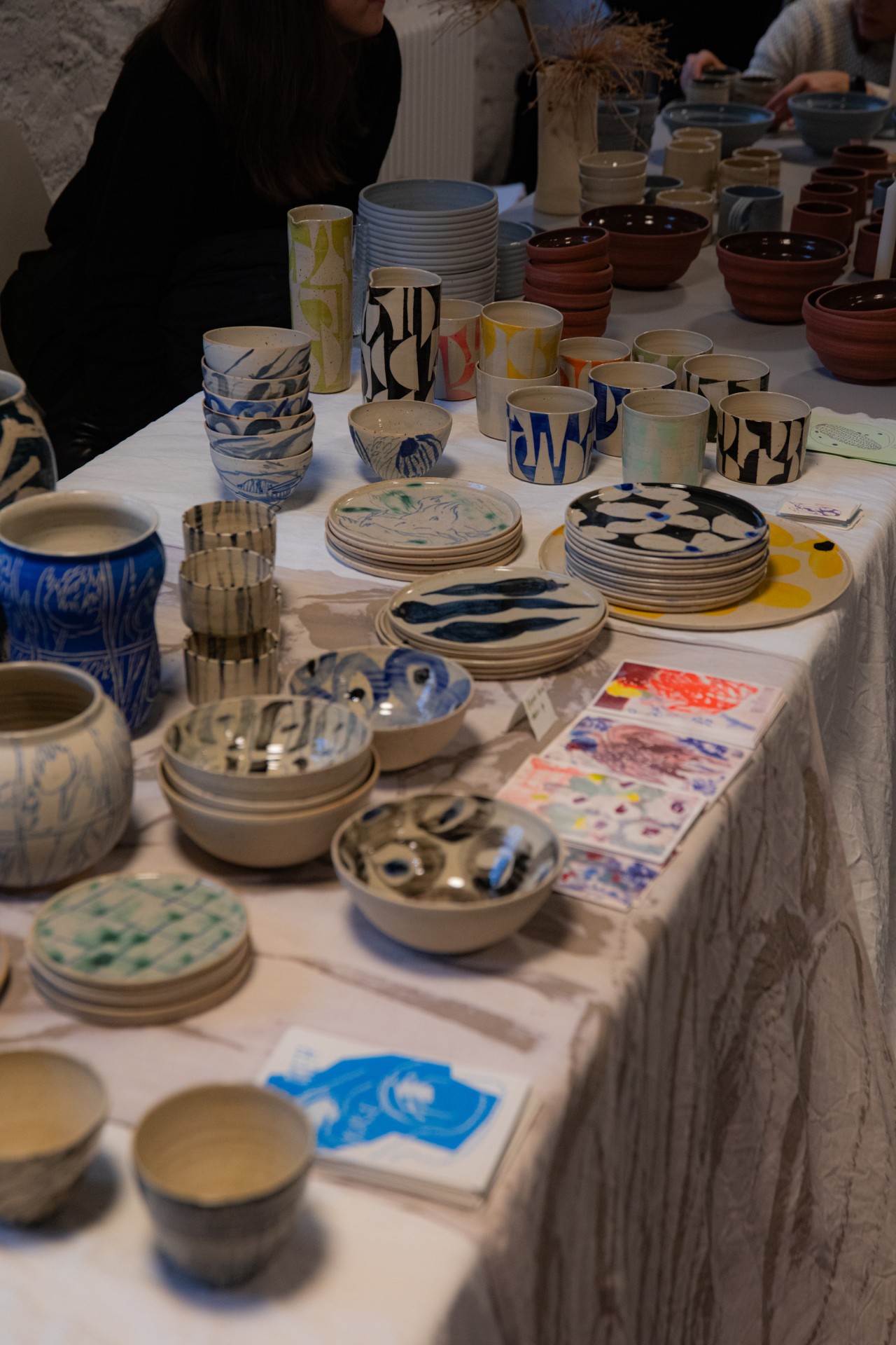 Tisch mit bunten Keramiktöpfen, Schalen und Tellern, umgeben von Menschen in einem kreativen Ambiente.