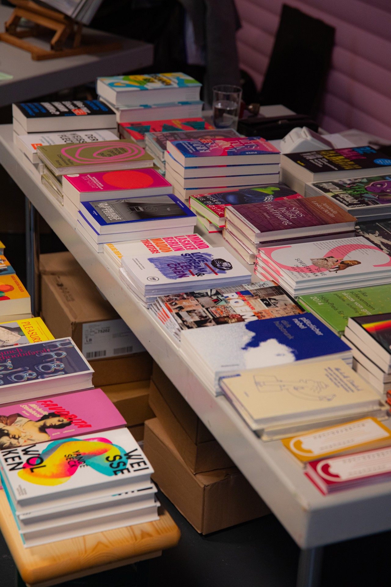 Tisch mit bunten Büchern und Notizen, umgeben von Kisten. Ein Glas Wasser steht ebenfalls darauf.
