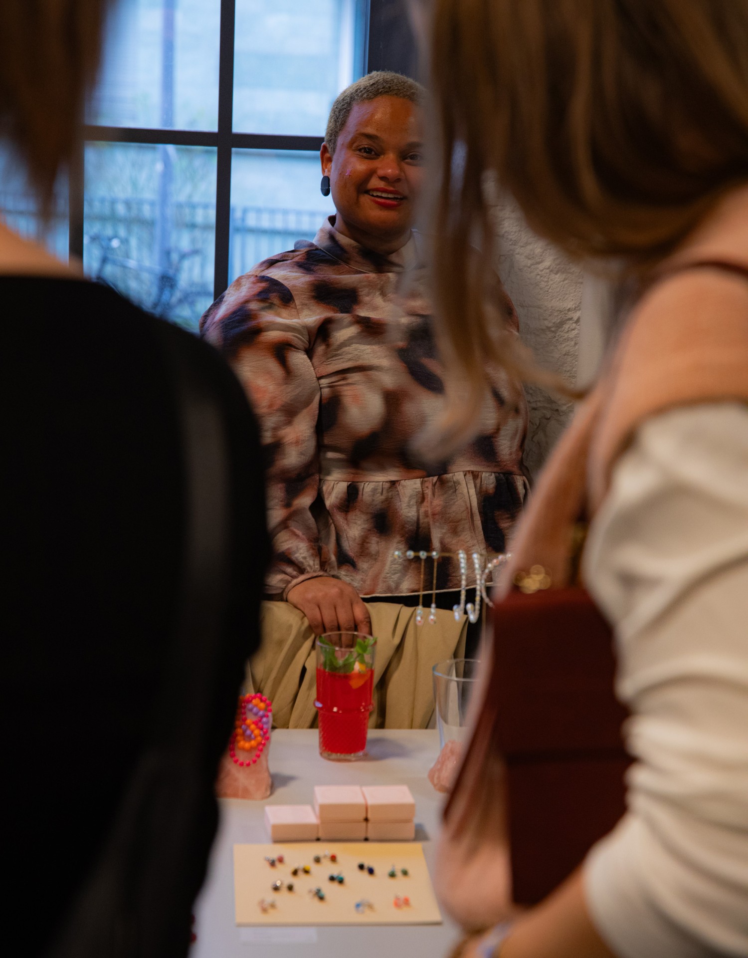 Eine Person in bunten Kleidern lächelt, umgeben von Gästen an einem Tisch mit Schmuck und Getränken.