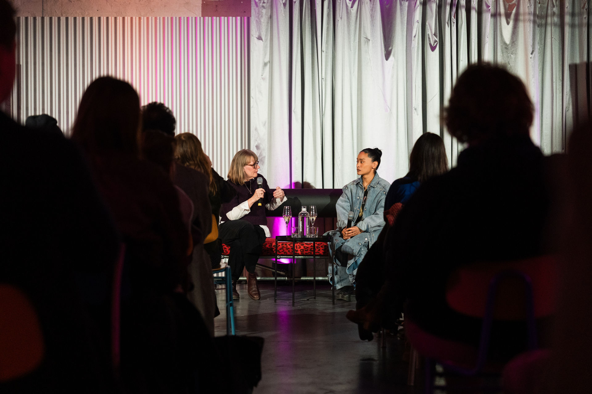 Zwei Frauen diskutieren auf einer Bühne, während das Publikum aufmerksam zuschaut. Eine professionell wirkende Atmosphäre.