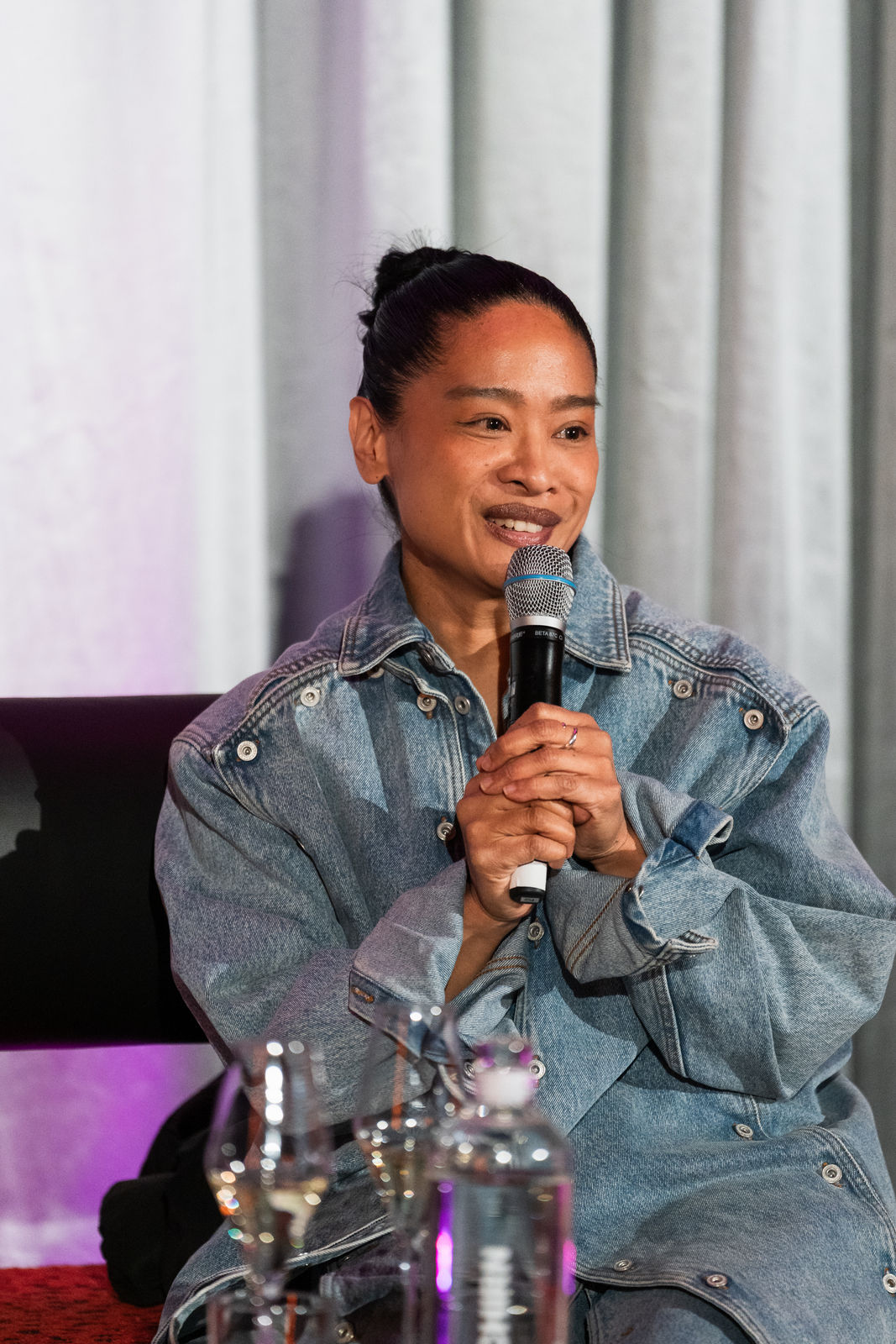 A person in a denim jacket speaks into a microphone, surrounded by glasses and water bottles on a table.