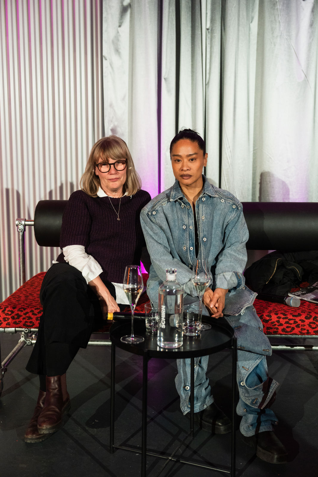 Zwei Frauen sitzen auf einem Sofa mit Gläsern und einem Tisch, hinter ihnen eine Vorhangstruktur in sanften Farben.
