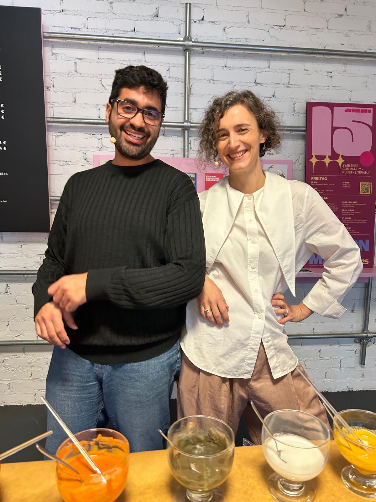 Zwei lächelnde Personen stehen an einem Tisch mit bunten Schüsseln vor einer weißen Backsteinwand.
