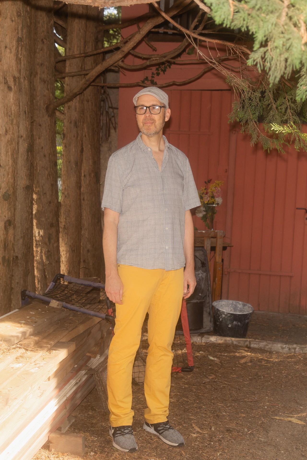 Mann in kariertem Hemd und gelben Hosen steht in einem Holzraum mit Bäumen und roter Wand im Hintergrund.