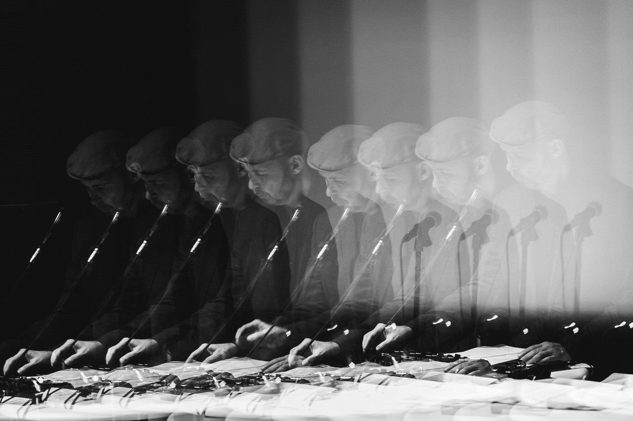 Ein Mann mit Schiebermütze spielt konzentriert an einem Tisch mit Mikrofonen in einem verschwommenen, künstlerischen Stil.