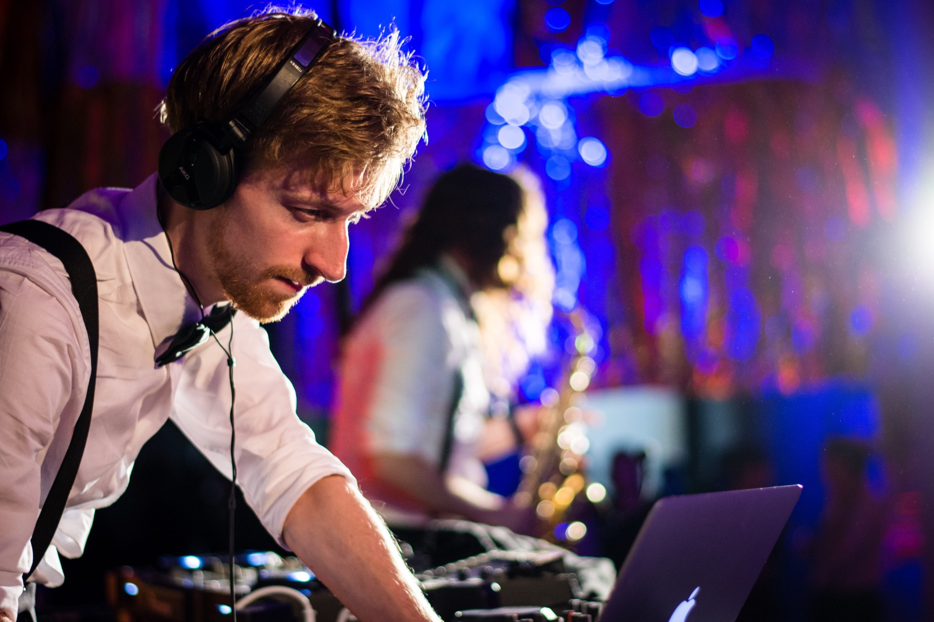 DJ with headphones at a mixing console, while a saxophonist performs in the background. Colorful lights create an atmosphere.