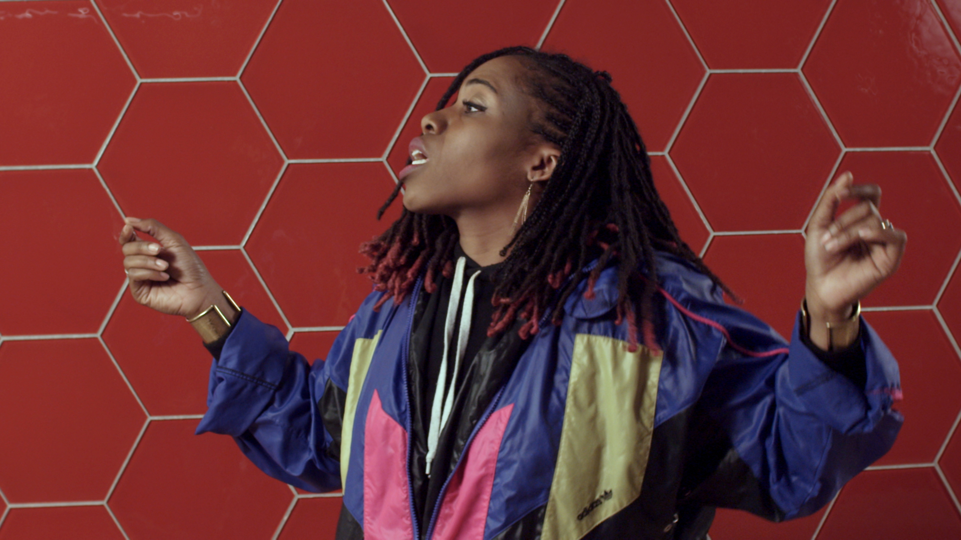 Eine Person mit Dreadlocks in bunter Sportjacke steht lebhaft vor einer rot gefliesten Wand.