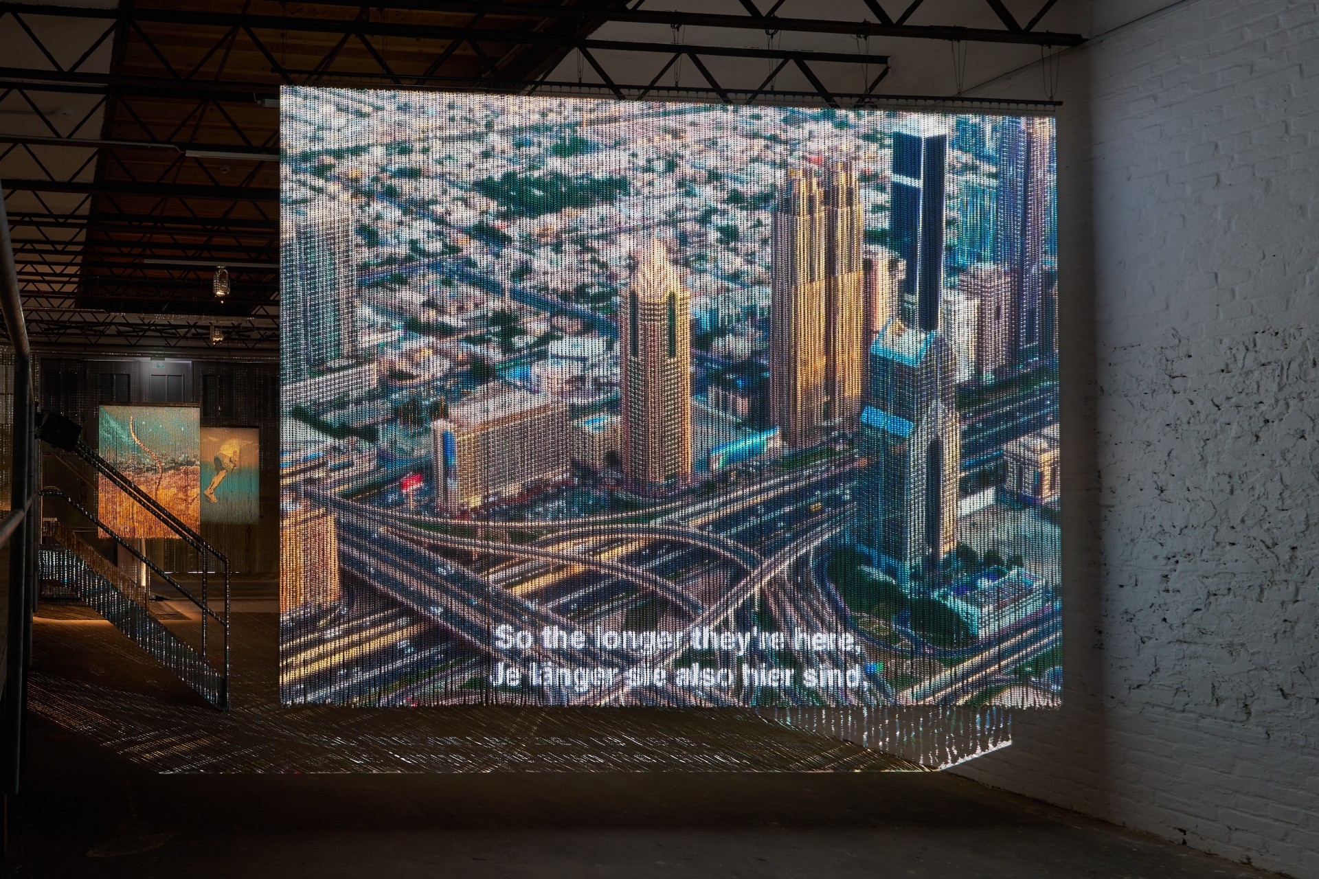 Installation eines Kunstwerks mit einem lebhaften Stadtbild auf einem Bildschirm, darunter Texte auf Englisch und Deutsch.