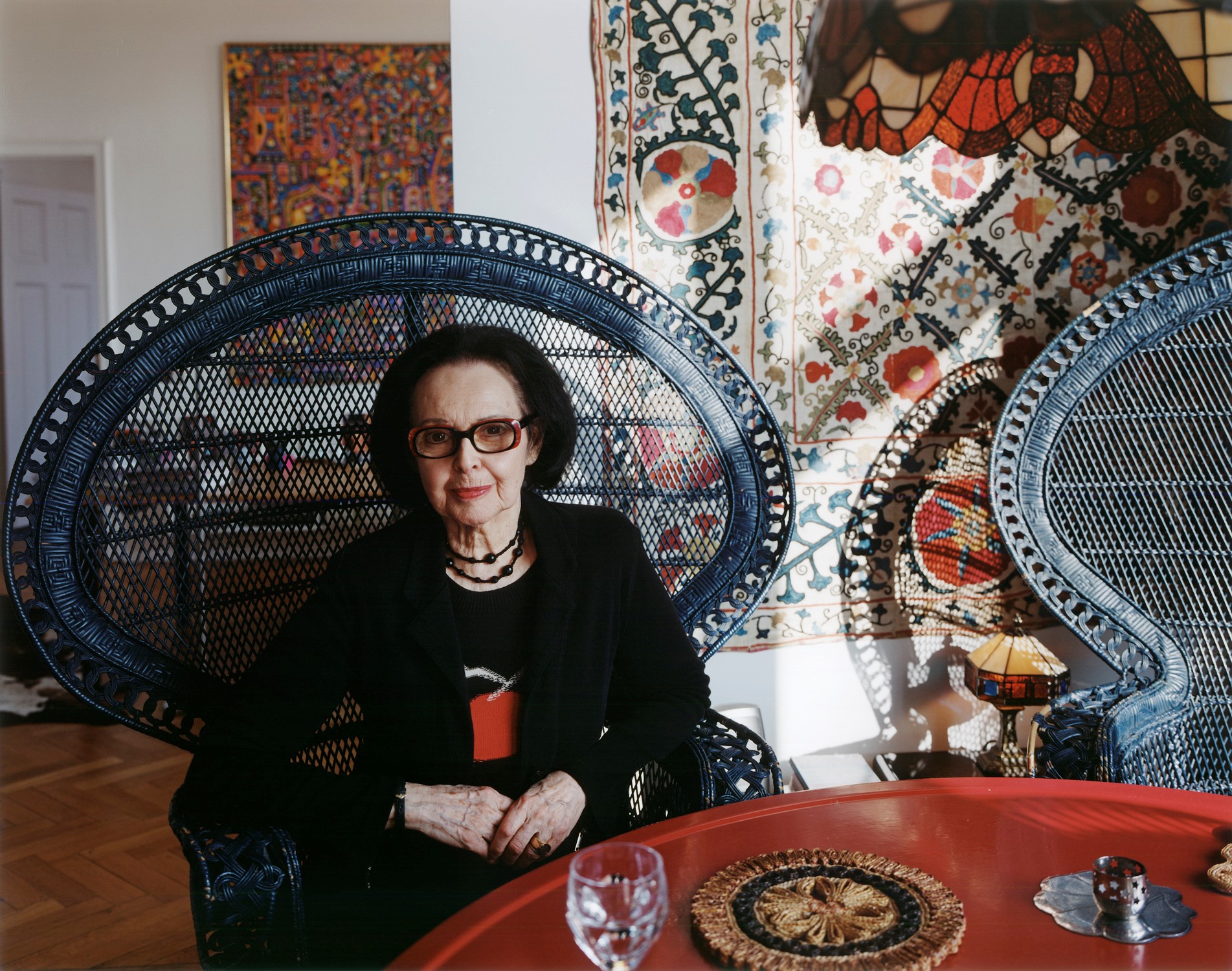 Ältere Frau in einem geflochtenen Stuhl, umgeben von bunten Textilien und Dekorationen, sitzt an einem roten Tisch.