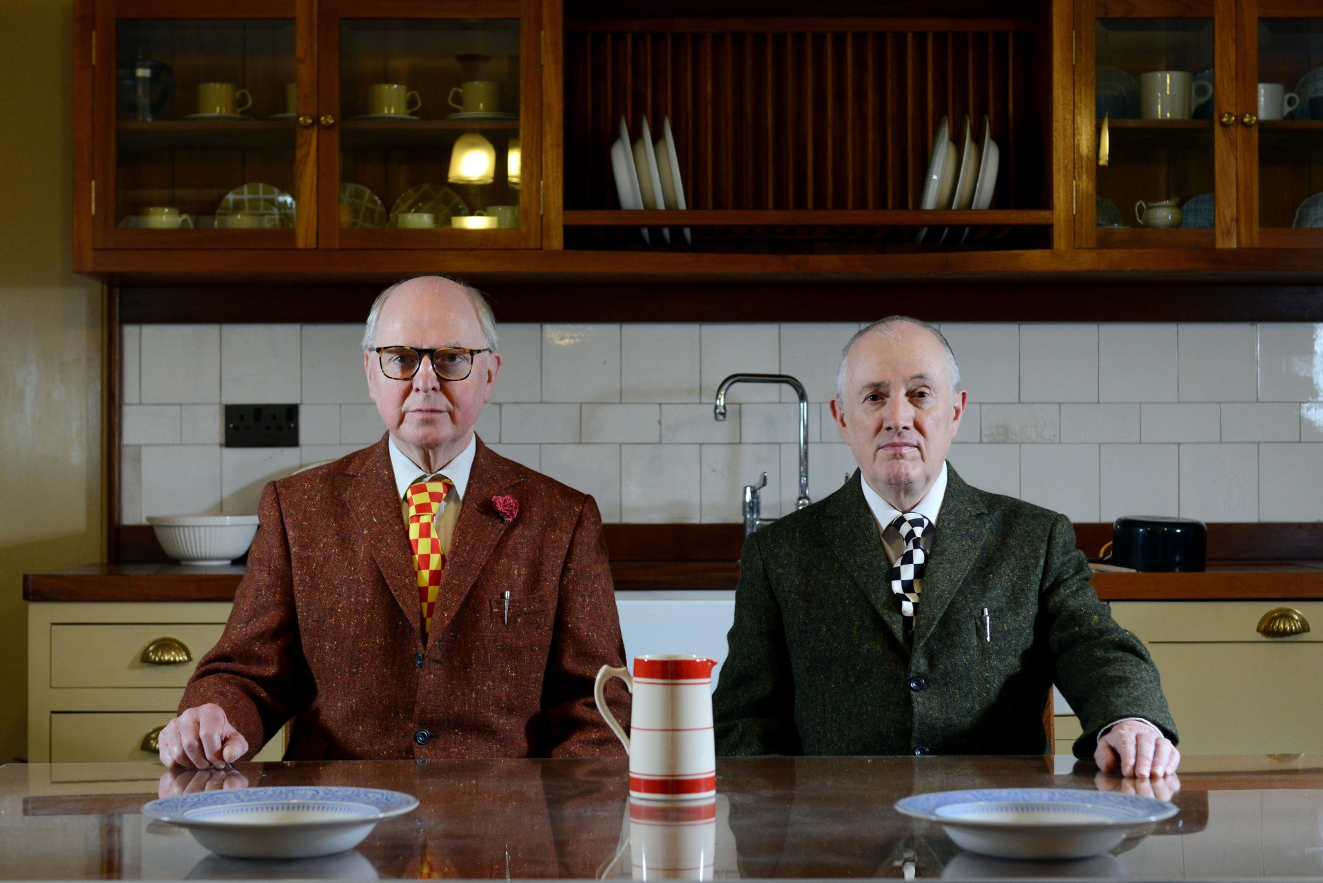 Zwei Männer in bunten Anzügen sitzen an einem Küchentisch, vor ihnen Geschirr und ein Krug.