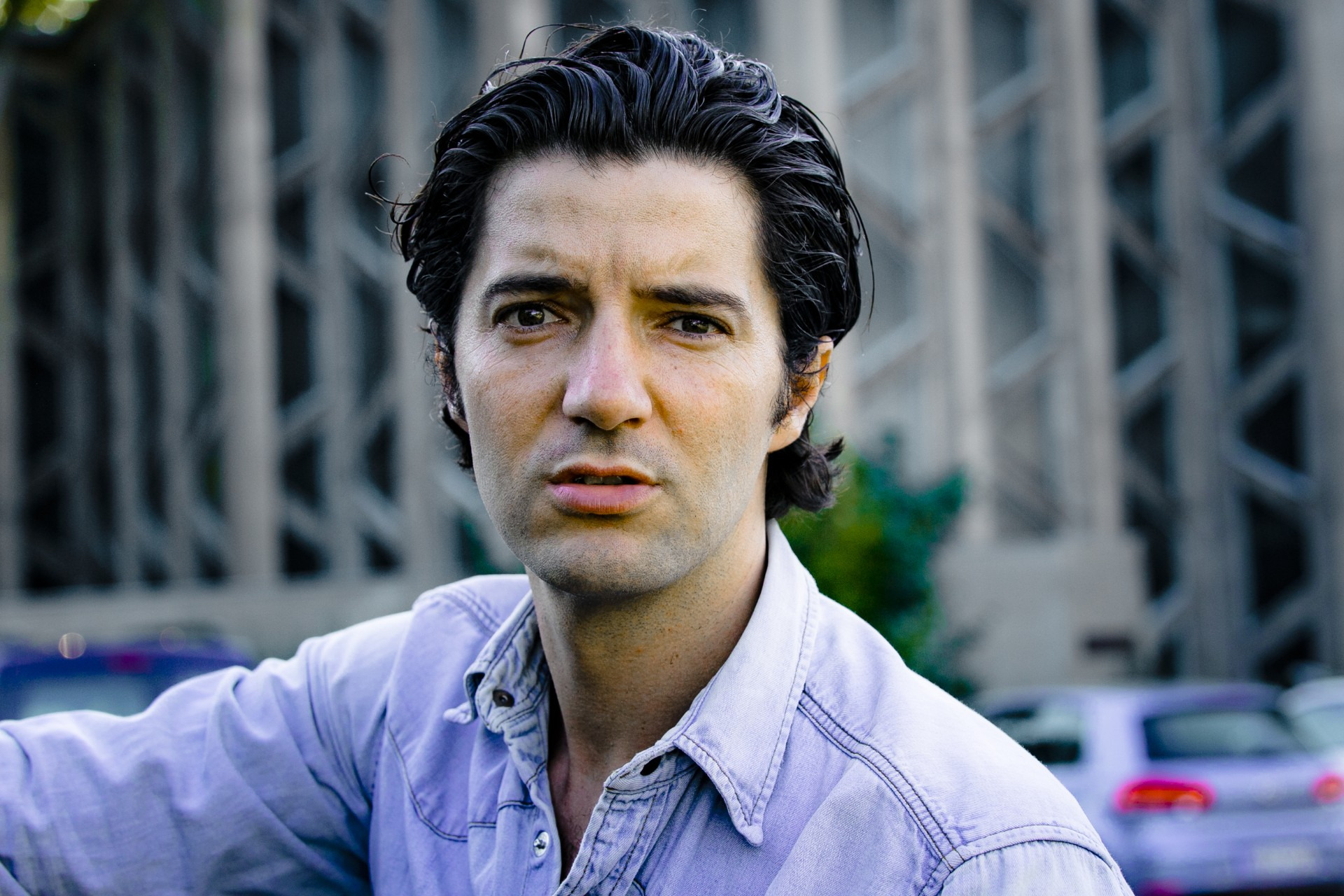 Ein Mann mit dunklen Haaren und ernstem Blick trägt ein helles Hemd vor einem modernen Gebäude.