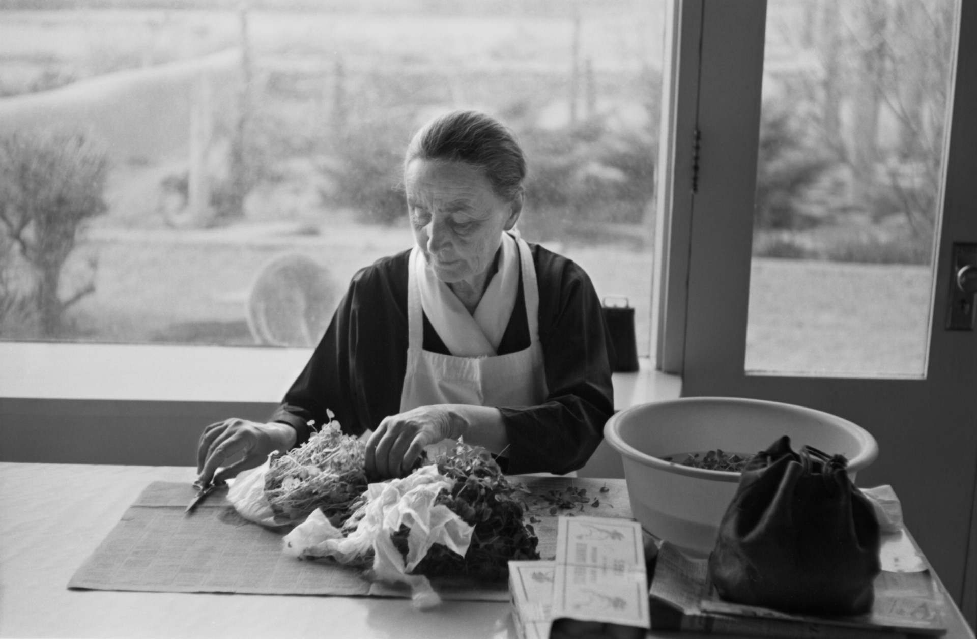 Ältere Frau in einem Raum, die sorgfältig mit Kräutern und Materialien arbeitet, von natürlichem Licht umgeben.